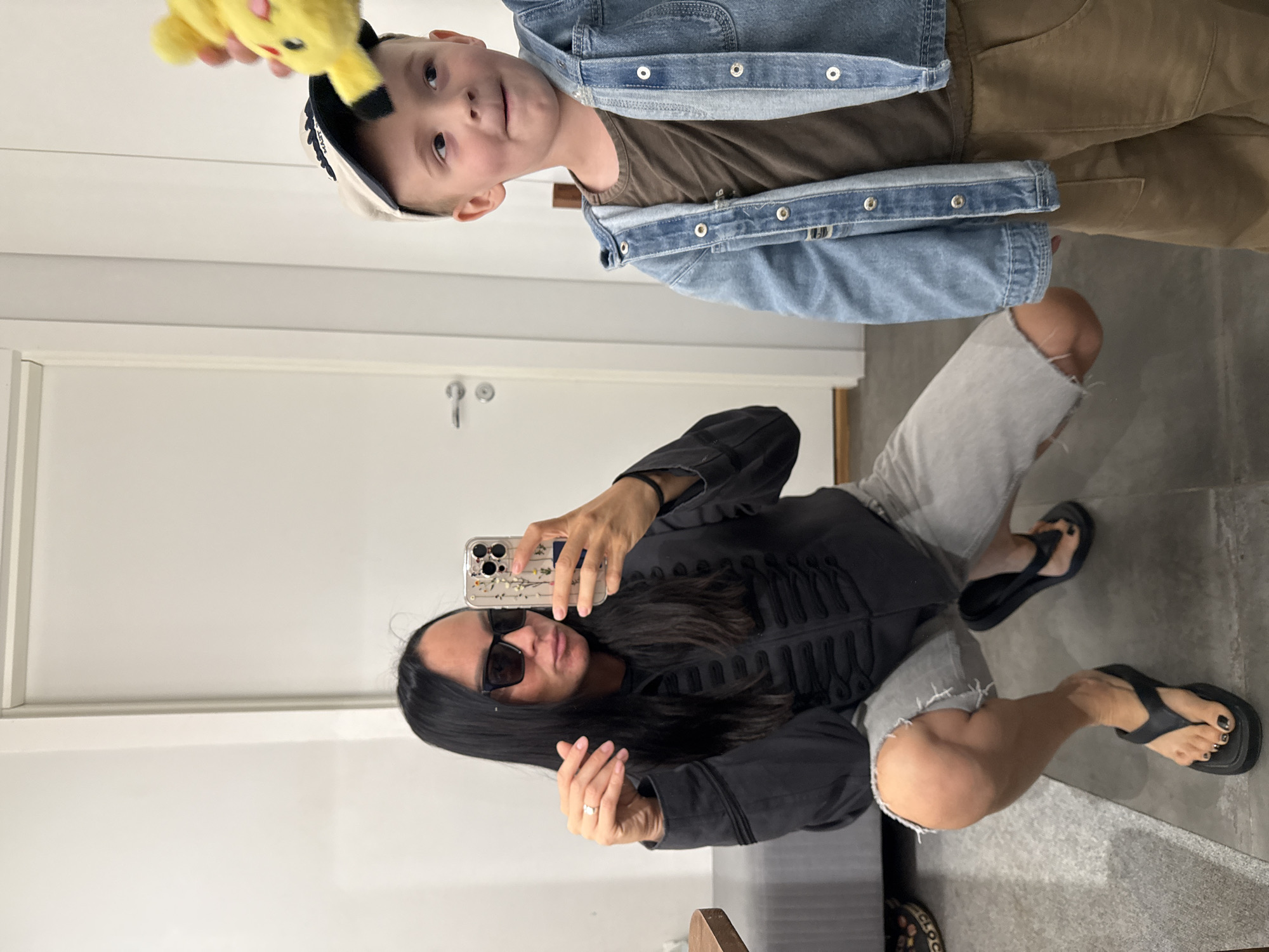 A woman and a young boy pose for a mirror selfie in an indoor setting that appears to be a domestic environment, possibly an apartment or house, in Chaikovsky, Russia. The woman, on the left, is squatting and wearing a dark-colored jacket with decorative buttons, light grey ripped denim shorts, and black flip-flops. She has dark hair and is wearing black sunglasses, and she is holding up her phone to take the picture. The boy, on the right, is standing and wearing a denim jacket over a brown t-shirt and tan pants. He has a light-colored baseball cap on and is looking towards the camera, with a yellow plush toy, possibly Pikachu, being held up by someone's hand above his head. The background shows a white door with a silver handle and a white wall. The floor is tiled.