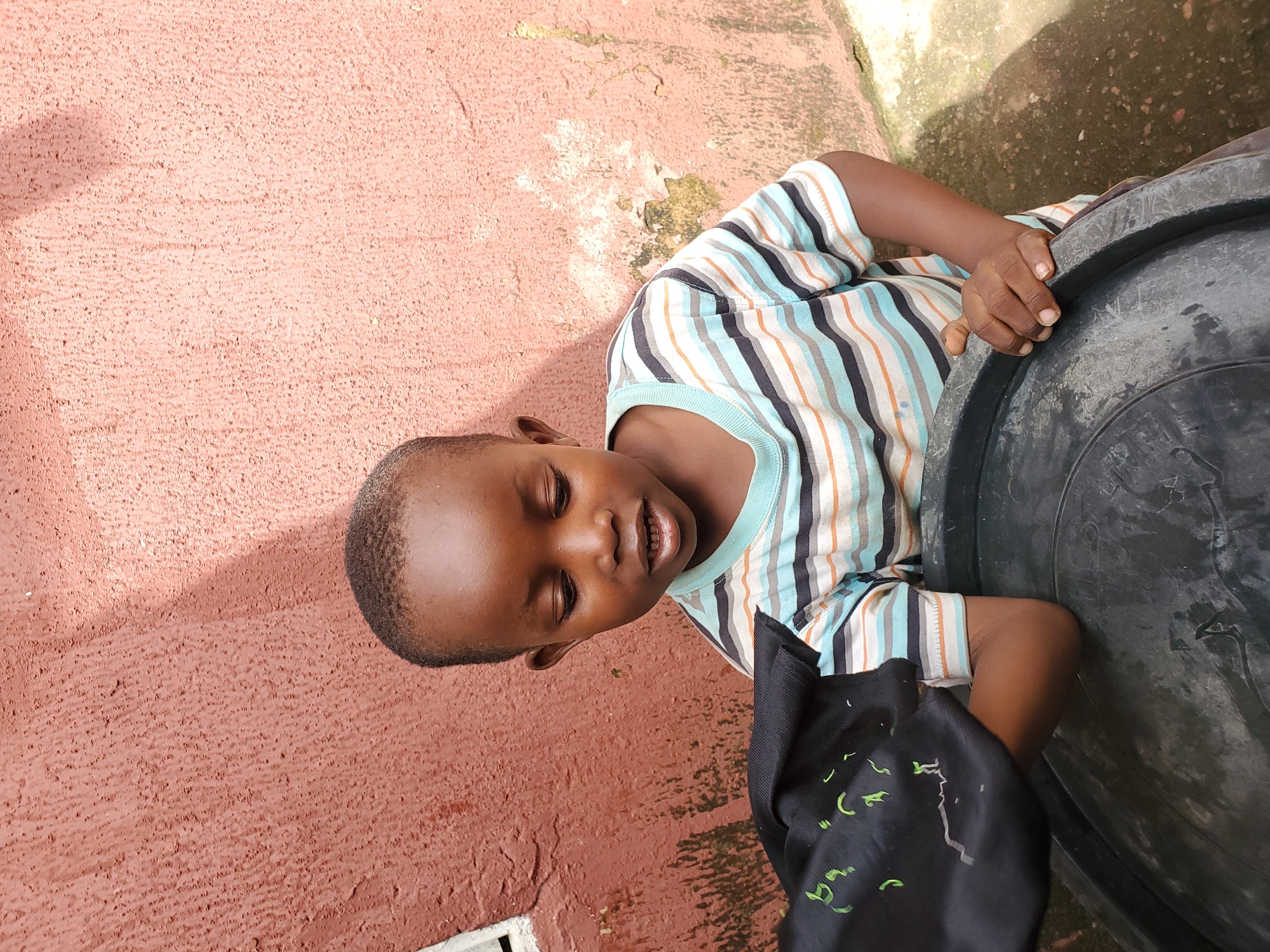 A young Black boy stands in front of a textured, reddish-brown wall with some water stains. He is wearing a horizontally striped t-shirt with white, blue, black, and orange stripes. He is holding a black fabric with green markings in his left arm and is resting his right hand on a large, black, round container. His gaze is directed downwards and he appears to be speaking. The scene is outdoors, with dappled sunlight creating shadows on the wall. The ground beneath the boy and the container appears to be paved and slightly wet. The image appears to have been taken in a residential or public outdoor area within the city of Jos, Nigeria, suggested by the architectural style of the wall and the ambient lighting.