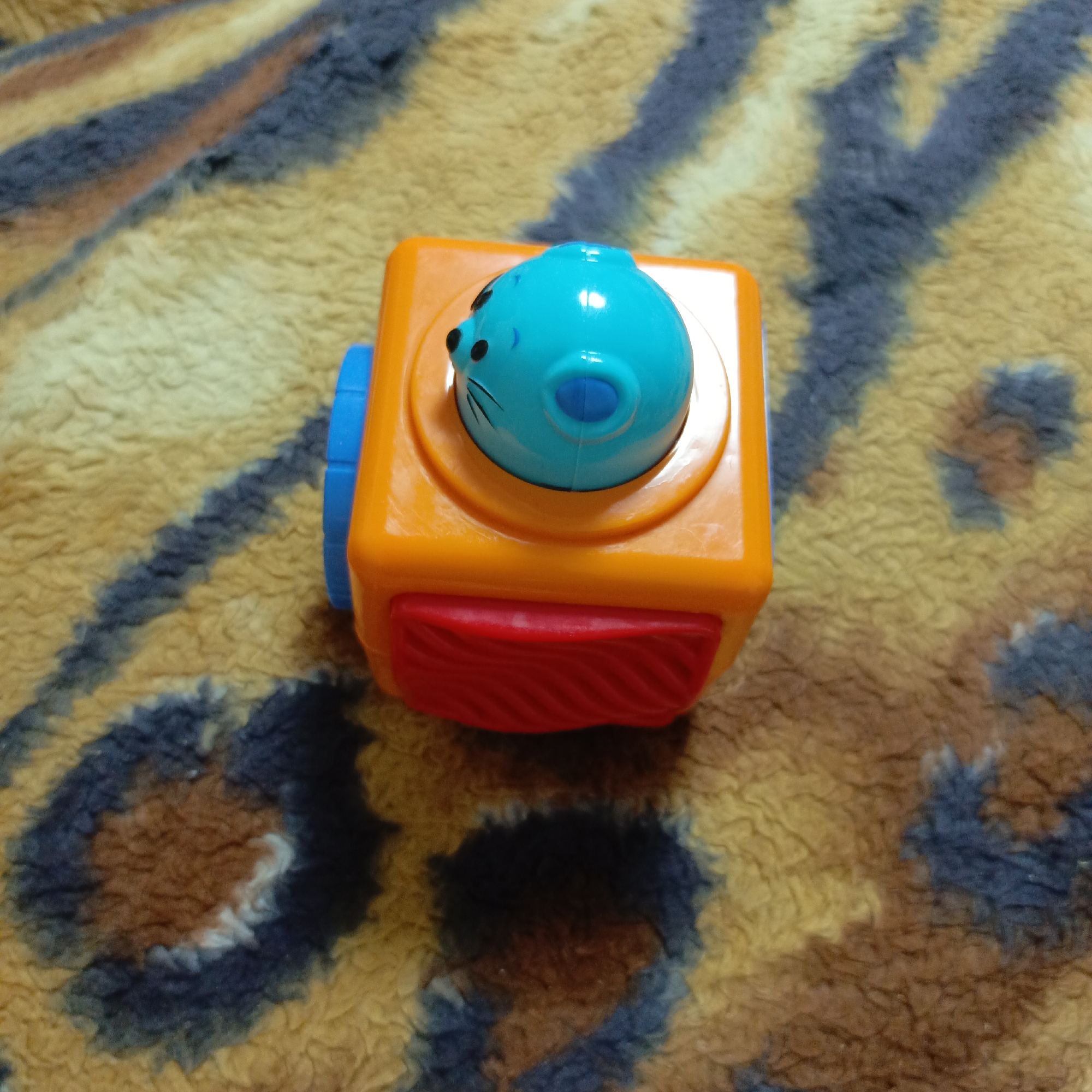 An overhead, slightly angled shot captures a colorful toy resting on a textured, patterned surface. The main subject is an orange, cuboid-shaped pop-up toy. On top of the orange base is a smooth, blue, rounded component, resembling an animal's head, possibly a seal or cat. This blue head has black dots for eyes and thin black lines depicting whiskers on its left side. A smaller, darker blue circular detail is also visible on the front of the blue head.
On the left side of the orange cube, a blue, ribbed cylindrical piece is attached. The front face of the orange cube features a red, rectangular, textured panel or button that appears to be part of the toy's interactive mechanism. The toy's plastic surface reflects some light, indicating a smooth, glossy finish.
The toy is situated on a soft, plush background, likely a blanket or rug, displaying an abstract, mottled pattern in shades of yellow, brown, and dark blue or grey. The varied colors and fuzzy texture of the background create a visually soft and warm setting. No activity is taking place, and the toy is static. There is no visible text in the image. The lighting appears even, suggesting an indoor setting.