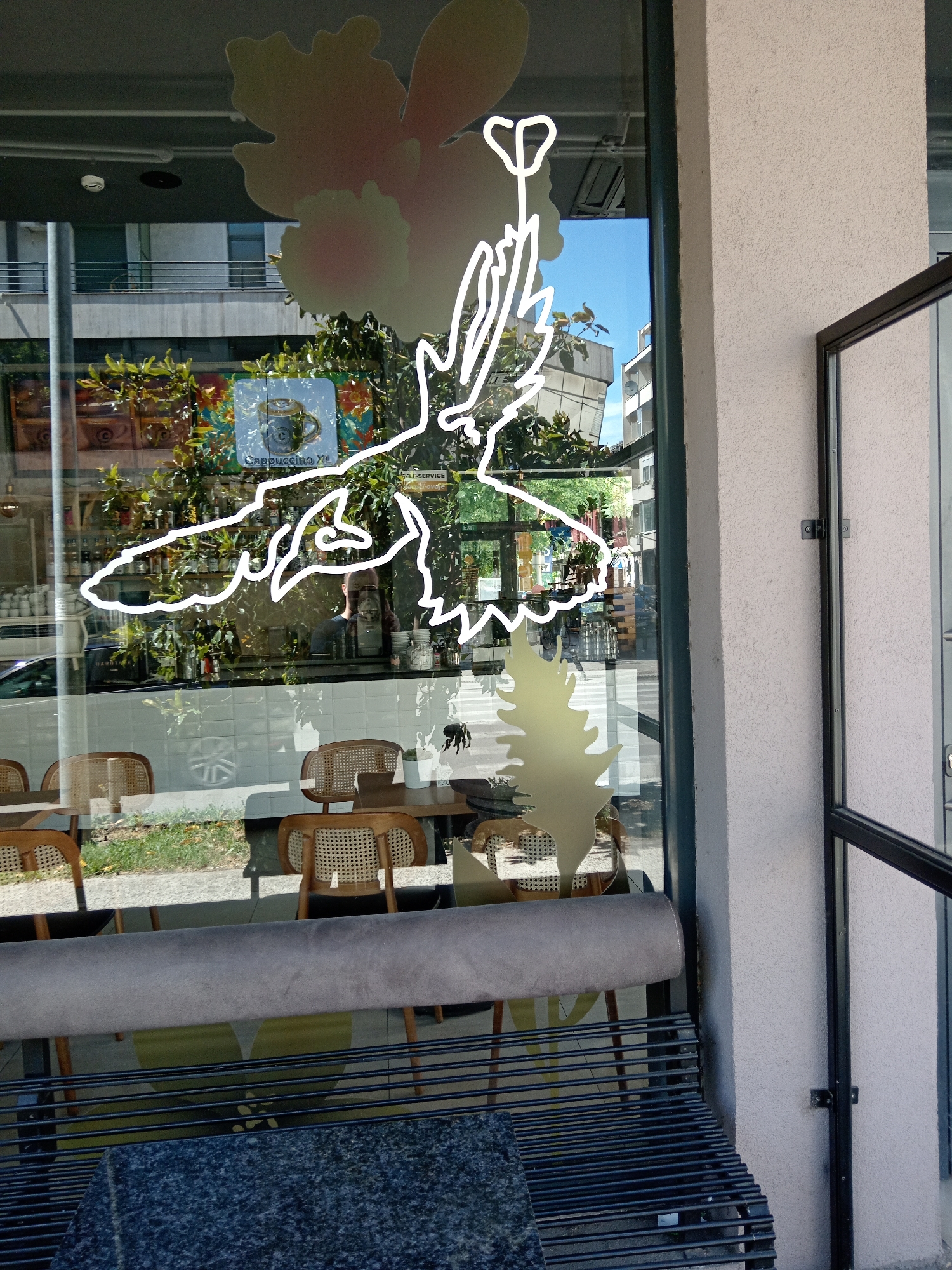The image presents a sunlit street view in Podgorica, Montenegro, framed by the large glass window of a cafe or shop. A prominent white, neon-like outline of a winged figure, possibly an angel holding a staff, is affixed to the window. Below this, decorative frosted decals depicting flowers and a stylized tree are also visible on the glass.
Through the window, the interior of the establishment shows wooden tables with chairs featuring woven cane backs. A long, grey upholstered bench runs along the inside, partially obscuring the interior furniture. In the immediate foreground, a black metal bench with a mottled grey, granite-like surface on top is positioned outside. Inside, behind a white-tiled counter, shelves hold various items like bottles and cups. A square sign with a coffee cup graphic and the text "Cappuccino Yu" is visible through the window. Faint text indicating "SELF-SERVICE" and an "EXIT" sign can also be seen.
Reflections on the glass reveal the urban surroundings: buildings across the street, lush green trees, and glimpses of a parked car and a pedestrian crosswalk. The bright light suggests a clear, sunny day. A dark, shadowy reflection of a person, likely the photographer, is faintly visible on the right side of the window. The scene captures a typical storefront on a city street during daylight hours.