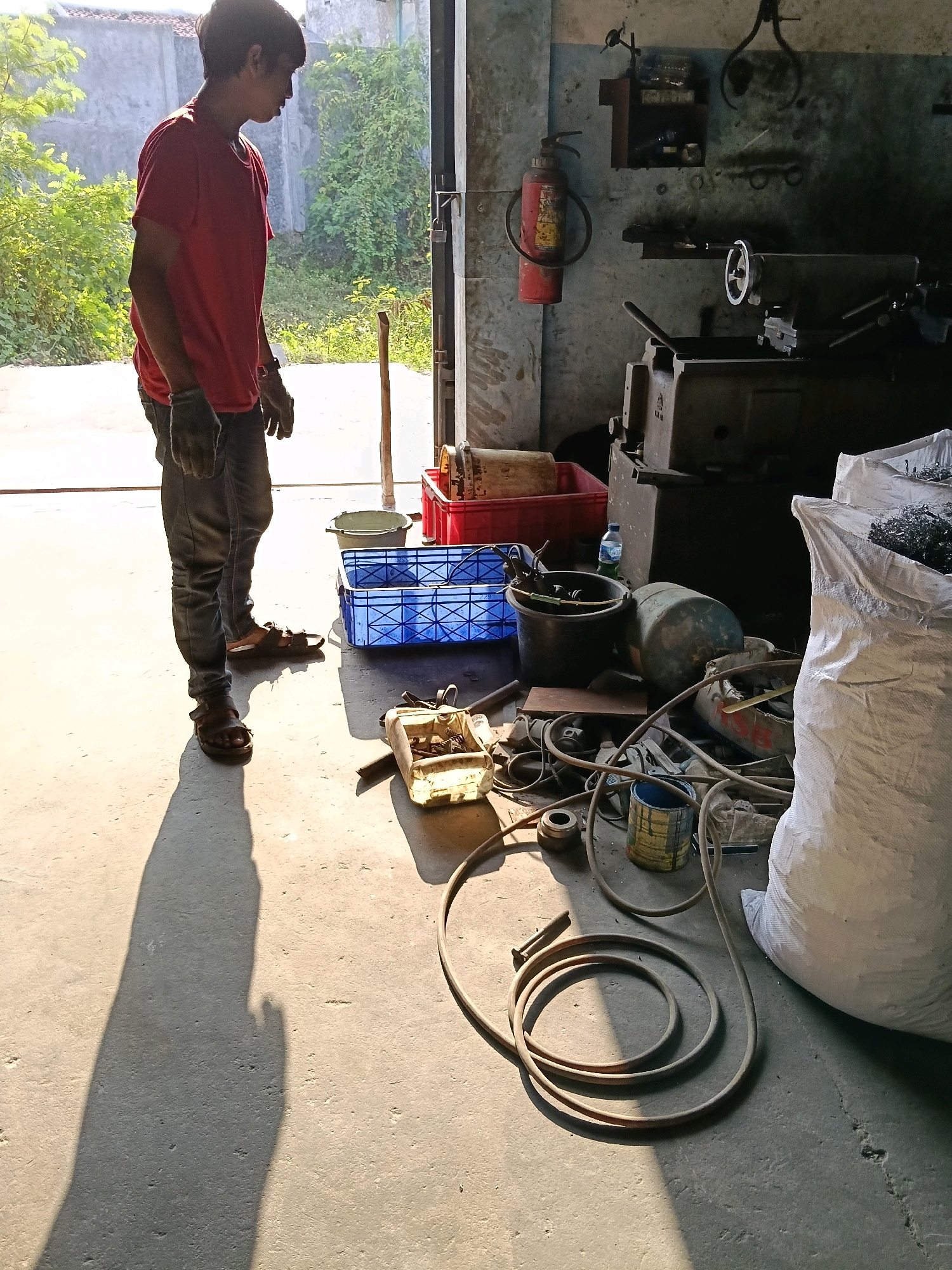 The image depicts a workshop interior, with sunlight streaming in from an open doorway, casting long shadows across the concrete floor. A young man, wearing a red t-shirt, grey jeans, and dark gloves, stands facing away from the camera, looking towards the left. He is positioned near the entrance, with lush green foliage visible outside.
The workshop is filled with various tools and equipment. To the right of the man, a fire extinguisher is mounted on the wall. A metal lathe is prominent in the background, alongside other industrial machinery. Various containers, including red and blue crates, are scattered on the floor, some containing materials. Several hoses and coiled cables are also visible, as is a large white sack, likely filled with scrap metal or other industrial waste. A small plastic water bottle sits on the floor near a dark bucket. The overall impression is of an active, albeit somewhat cluttered, workspace. The setting appears to be a workshop in Belimbing, Indonesia, suggested by the context provided.