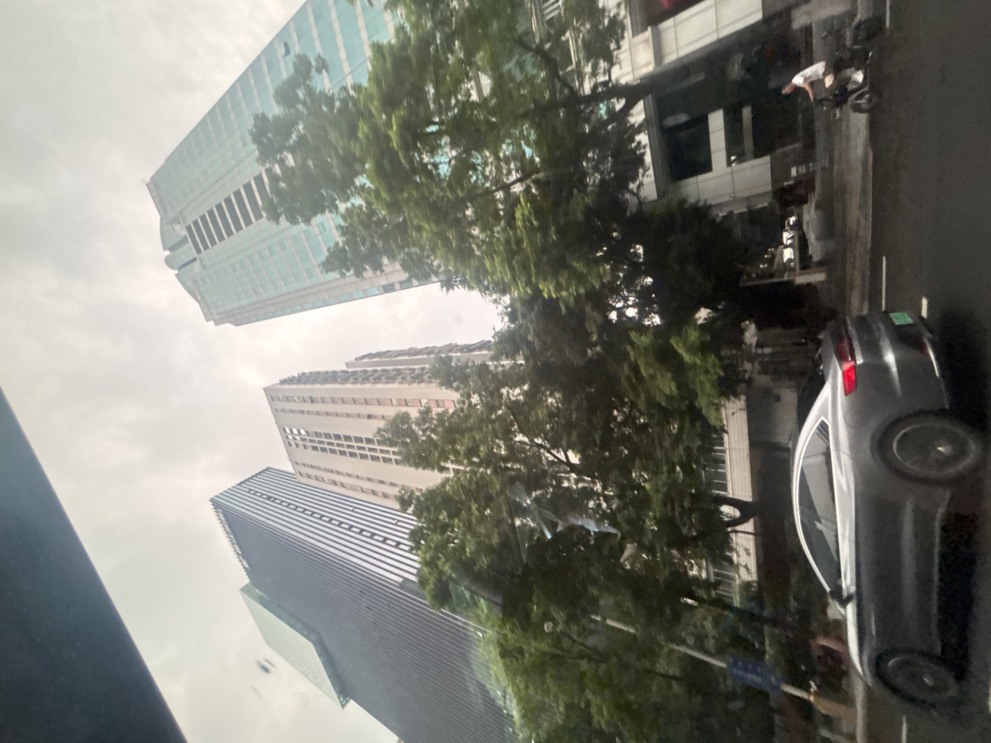 An eye-level, medium outdoor shot taken from a vehicle shows a gray electric sedan driving on a wet street in Yuexiu District, China. The sky is overcast. Buildings rise behind the street, including a modern glass-fronted skyscraper with a teal tint, a darker skyscraper with a striped facade, and a lighter-colored building with many windows. Trees line the street in front of the buildings. A person is riding a small electric scooter on the street to the right of the sedan.