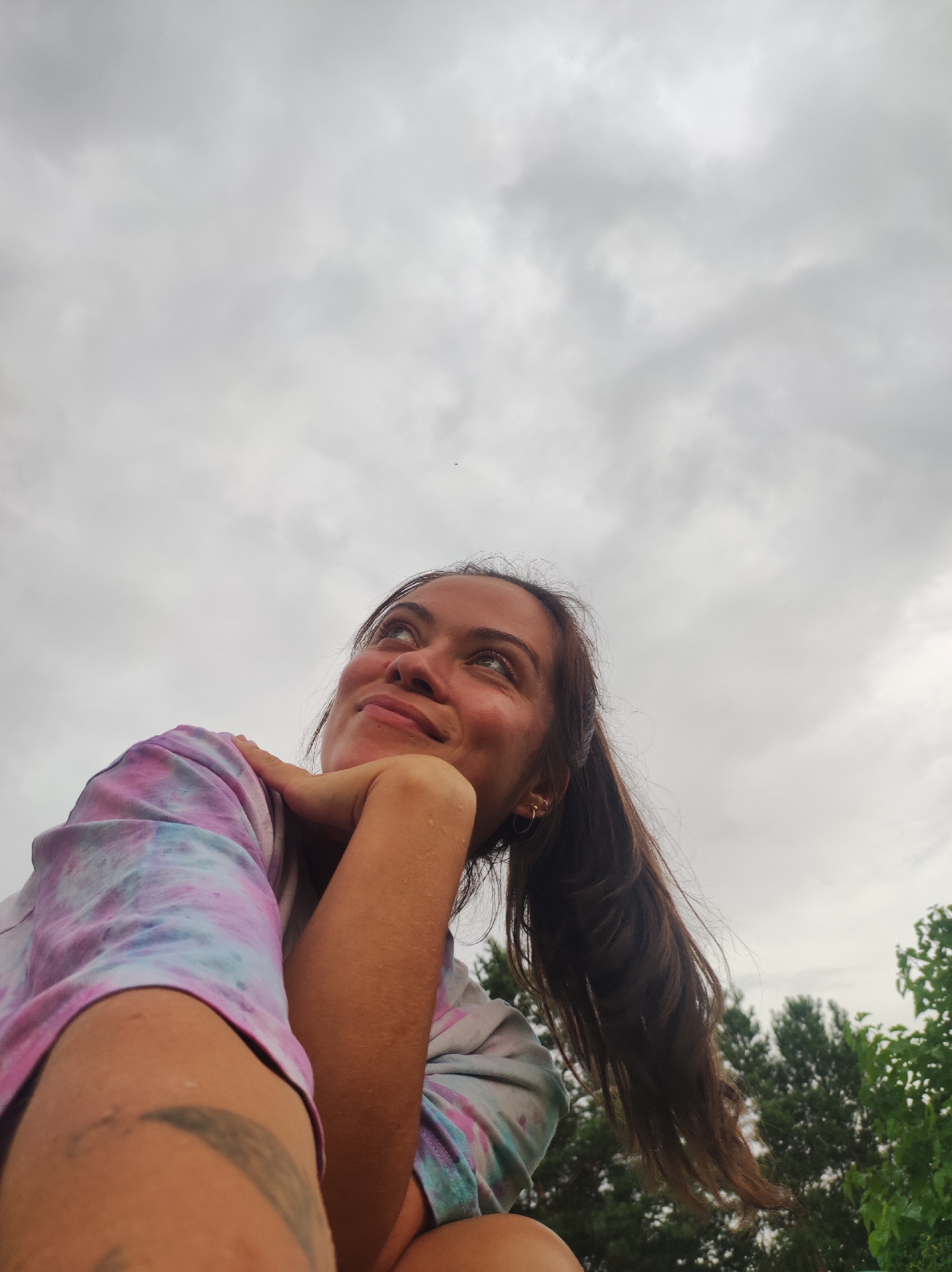 A low-angle, medium shot captures a young woman from the chest up against a cloudy sky. She is looking upwards and to her left with a smile on her face, her left arm bent with her hand resting on her chin. Her right arm is bent at the elbow, with her hand resting on her shoulder. She is wearing a tie-dye t-shirt with pink, purple, and blue colors. Her long brown hair is pulled back in a ponytail. In the background, trees are visible at the bottom right of the frame. The scene appears to be outdoors, with no specific indicators of the city of Фоки, Russia.