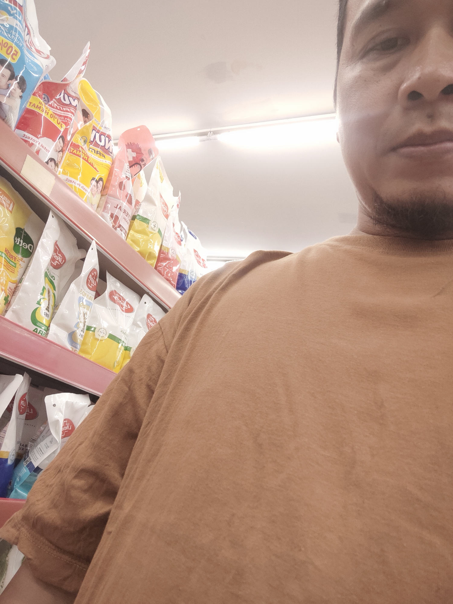 The image is a close-up, slightly tilted, first-person perspective of a person in a supermarket in Desa Banyumas, Indonesia. The person is wearing a brown t-shirt, and the right side of their face, including their eye, nose, and mouth, is visible. The background shows shelves stocked with various packaged goods, primarily laundry detergent or similar cleaning products, with colorful branding and images of people. The lighting suggests an indoor environment with fluorescent overhead lights. The scene is static and focuses on the individual's presence in the retail setting.