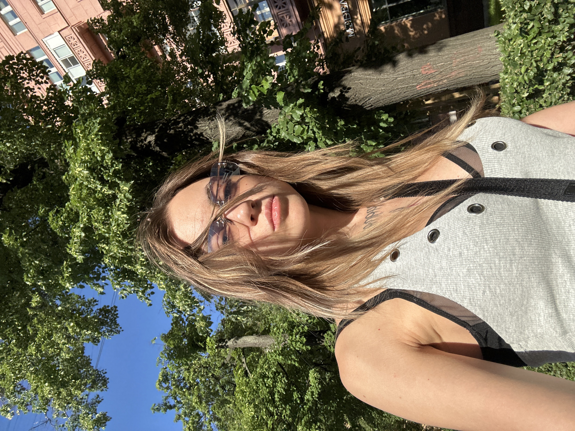 An outdoor, low-angle portrait depicts a fair-skinned female, approximately 20-35 years old, looking directly at the camera. Her medium-length, light brown hair, styled with blonde highlights, is windblown across her face. She wears thin-framed sunglasses with dark lenses. Visible tattoos, appearing as dark script or abstract designs, are present on her upper chest/neck area. She is dressed in a light grey ribbed tank top featuring metallic grommets along the front edges, under which a black mesh bra or bralette is visible. A black strap, likely from a bag, crosses diagonally over her chest. Her right shoulder and arm are exposed.
The background consists of a clear blue sky, lush green deciduous trees, and their textured trunks, suggesting a park or tree-lined street under bright sunlight. To the right, a multi-story building with a light pink/peach facade is prominent. It features multiple windows with white frames and a distinctive balcony with geometric patterns on a higher floor. Partially visible Cyrillic text, reading "МОСКВА" (MOSCOW), is present on the building's facade, confirming the location as the city of Moscow, Russia. Green bushes or hedges line the foreground at the base of the building. The scene is illuminated by direct sunlight, creating strong highlights and shadows.