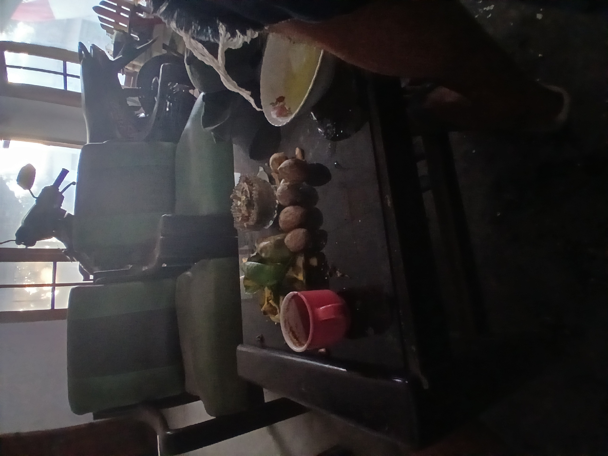 In a dimly lit interior, likely in Kademangan Kulon, Indonesia, a dark wooden table occupies the foreground. On the table, a small red mug filled with a dark liquid, possibly coffee, sits to the left. To the right of the mug, a scattering of nuts, some appearing to be walnuts, are visible alongside a small pile of what seem to be leafy green vegetables or herbs. Further to the right, a shallow bowl contains a light yellow liquid, possibly soup or broth, with some white floral-patterned detail on its rim. Behind the table, two dark green upholstered chairs are partially visible, with a motorcycle parked behind them. Sunlight streams through a window in the background, illuminating the scene and casting shadows. A person's legs, clad in dark pants, are visible on the right side of the image, suggesting their presence in the scene. The overall impression is of a casual, domestic setting.