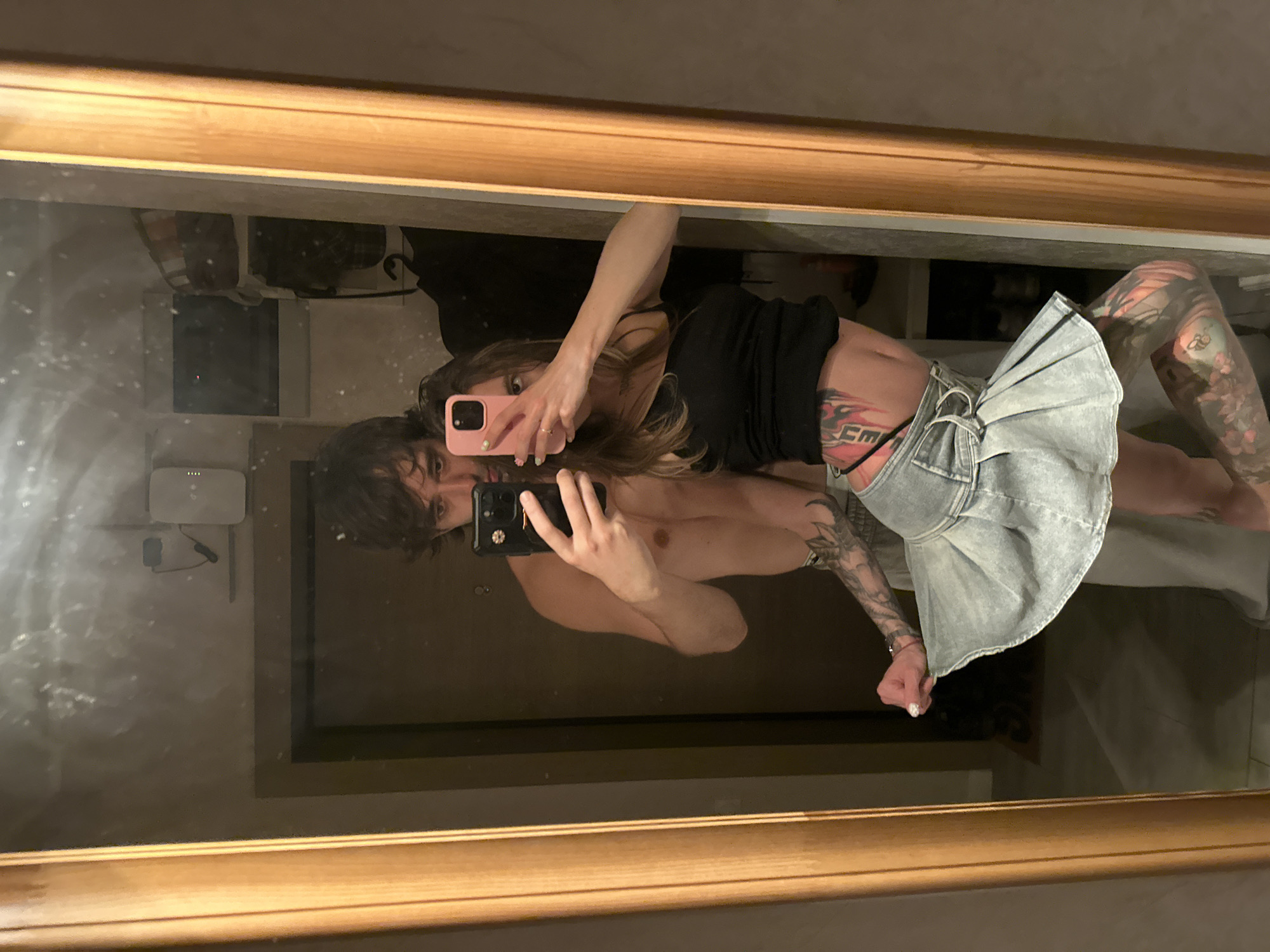 In a dimly lit room, a young couple takes a mirror selfie. The man, with dark, wavy hair, stands slightly behind the woman, holding a smartphone in his right hand. He is shirtless, and his left arm is around the woman. The woman, with long brown hair, faces the mirror directly, holding a pink smartphone in her right hand. She is wearing a black tank top and a light blue denim skirt. Her left hand is resting on her hip. Both individuals have visible tattoos. The background shows a closet filled with clothes and shelves. The room appears to be an apartment, possibly in Moscow, Russia, given the style of the furniture and decor.