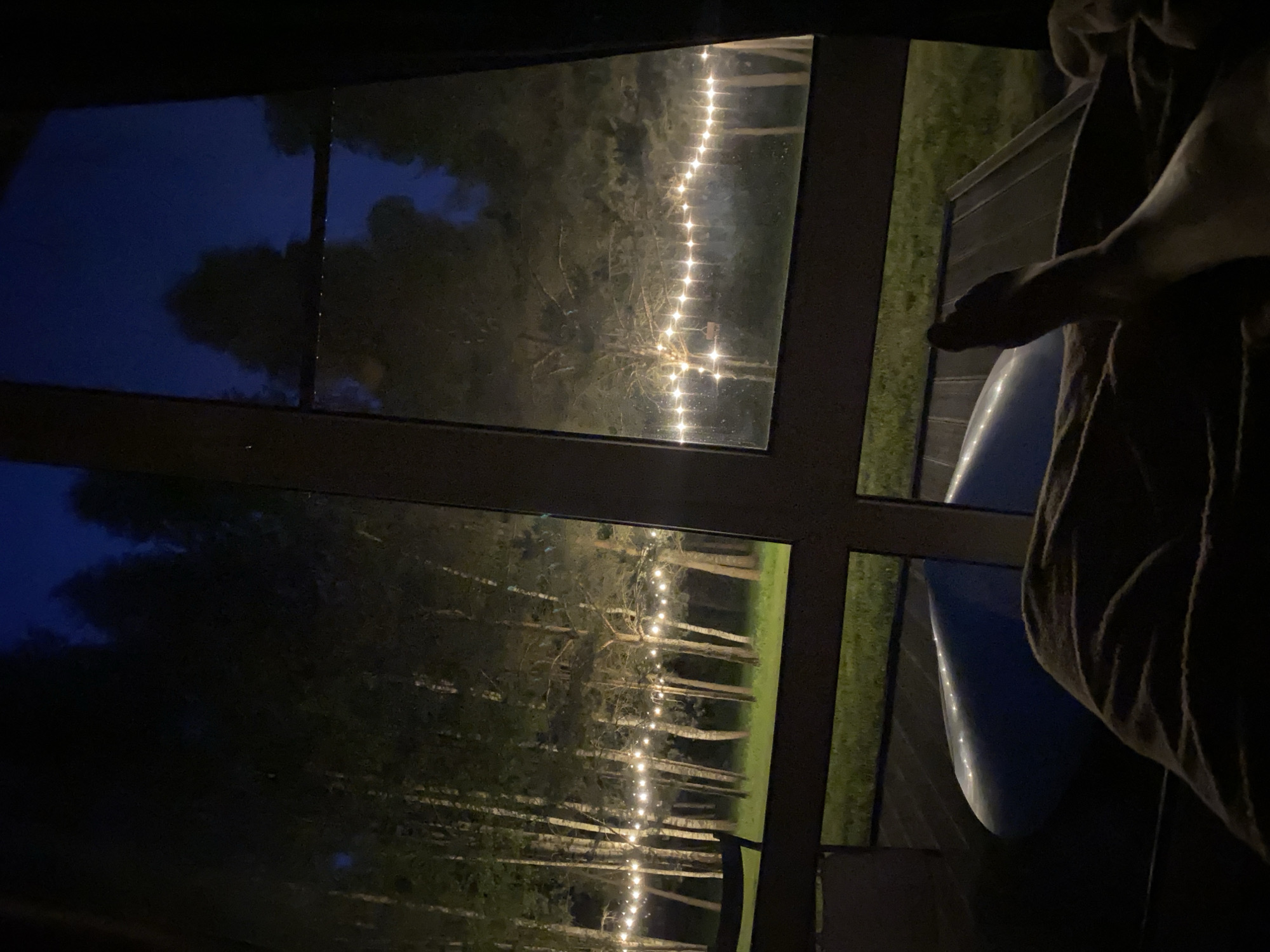 The image presents a night scene observed from indoors through a multi-paned window or glass door in Копнино, Russia. In the immediate foreground, a bare human lower right leg and foot are visible, resting on a dark, textured blanket or duvet. To the left, a smooth, light blue, curved object with a reflective surface is partially present on a dark floor or surface. The window's dark, thick frame is prominent, segmenting the view.
Outside, a dense line of trees, including slender birches with light bark and darker coniferous trees, stands against a deep blue, unlit night sky. A horizontal string of warm, glowing light bulbs is suspended between the trees at a uniform height, illuminating the tree trunks and the grassy ground below. A second, parallel string of lights is visible further back in the right pane. A manicured green lawn extends from the base of the trees. Immediately beyond the window frame, a dark wooden or composite deck surface is visible, with a portion of a dark outdoor chair or furniture piece positioned on the left side of the deck. Minute droplets, consistent with rain or condensation, are faintly visible on the window glass. A small, dark, rectangular object, possibly a bird feeder, is suspended from one of the trees in the right section of the view.