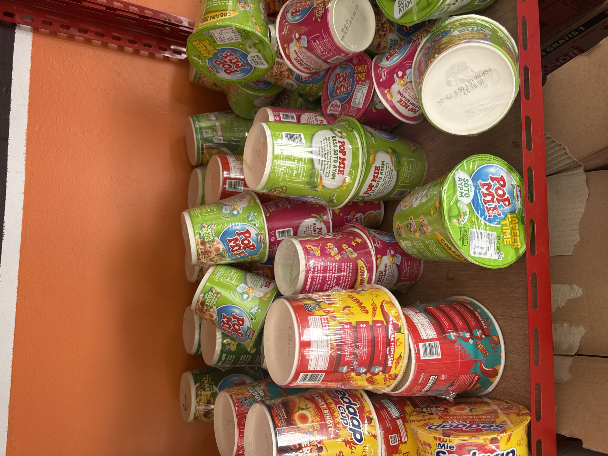 The image depicts a close-up, eye-level view of a shelf filled with numerous cups of instant noodles. The noodles are of various brands and flavors, predominantly "Pop Mie" and "Soto Ayam". Some cups are stacked vertically, while others are tilted. The overall color palette is dominated by green, red, and yellow from the packaging. The background is an orange wall with a white stripe at the top, and a red metal shelf unit is partially visible on the right. The shelf the noodles are on appears to be made of wood. The scene is well-lit, suggesting an indoor environment, possibly a retail store or a stockroom. There are no people visible in the image, and the focus is entirely on the product display. There is no indication of the specific location within Abang, Indonesia.
