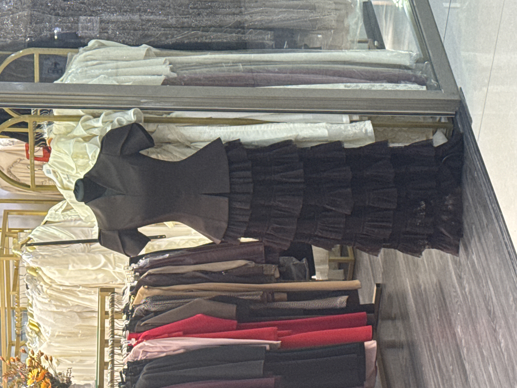 This is a medium-shot, eye-level photograph of a clothing store in Bishkek City, Kyrgyzstan. The image is dominated by a mannequin wearing a black, ruffled, floor-length dress. The dress features a high neckline, short puffed sleeves, and a cinched waist with a peplum detail. The skirt of the dress is composed of multiple tiers of ruffles.
To the left of the mannequin, racks of clothing are visible. These include a selection of brightly colored garments, such as red and pink dresses, as well as more muted tones like beige and grey. Further to the left, a vase with yellow and orange flowers can be seen.
Behind the mannequin and to the right, through a glass partition, additional garments are displayed on hangers. These appear to be lighter-colored, possibly white or cream, with ruffles and intricate detailing. The store's flooring is a light gray, wood-patterned laminate. The overall impression is one of an indoor boutique or retail space.