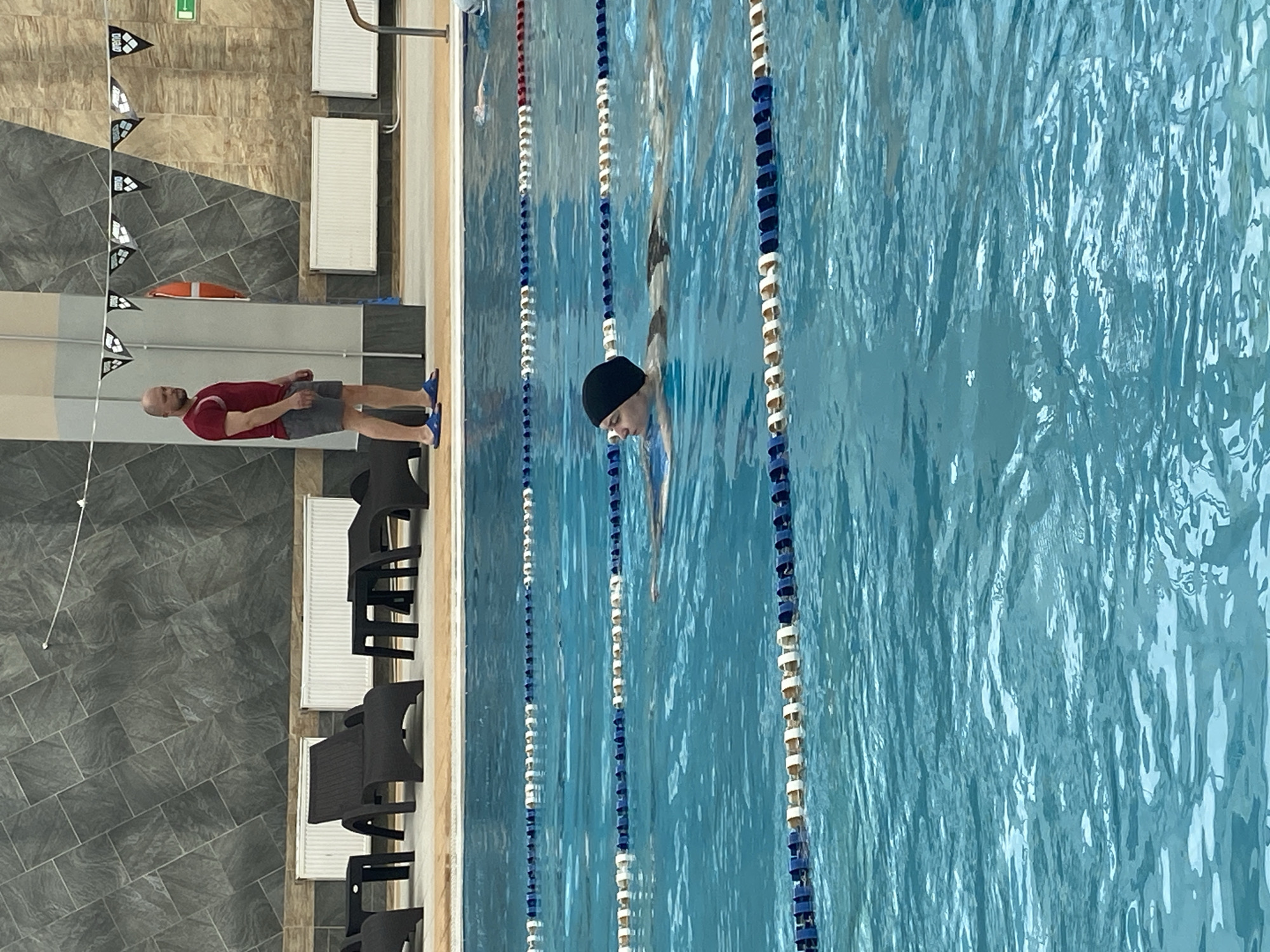 The image depicts an indoor swimming pool facility, likely located in Yekaterinburg, Russia, given the context. The pool water is a clear turquoise blue, divided into lanes by white and blue buoys. In the foreground, a person wearing a black swimming cap and dark swim trunks is actively swimming, holding a kickboard. Their face is partially visible, turned to the left.
In the background, on the pool deck, a man stands wearing a red t-shirt and grey shorts, with blue flip-flops. He appears to be observing the swimmers. Behind him, a wall is covered in dark, textured tiles. Further back, there are white radiators and several dark plastic lounge chairs and small tables arranged near the edge of the pool area. A life preserver is visible mounted on the wall. A string of triangular flags, some bearing the "arena" logo, hangs across the upper portion of the background. The lighting suggests an indoor environment, possibly a public swimming facility.