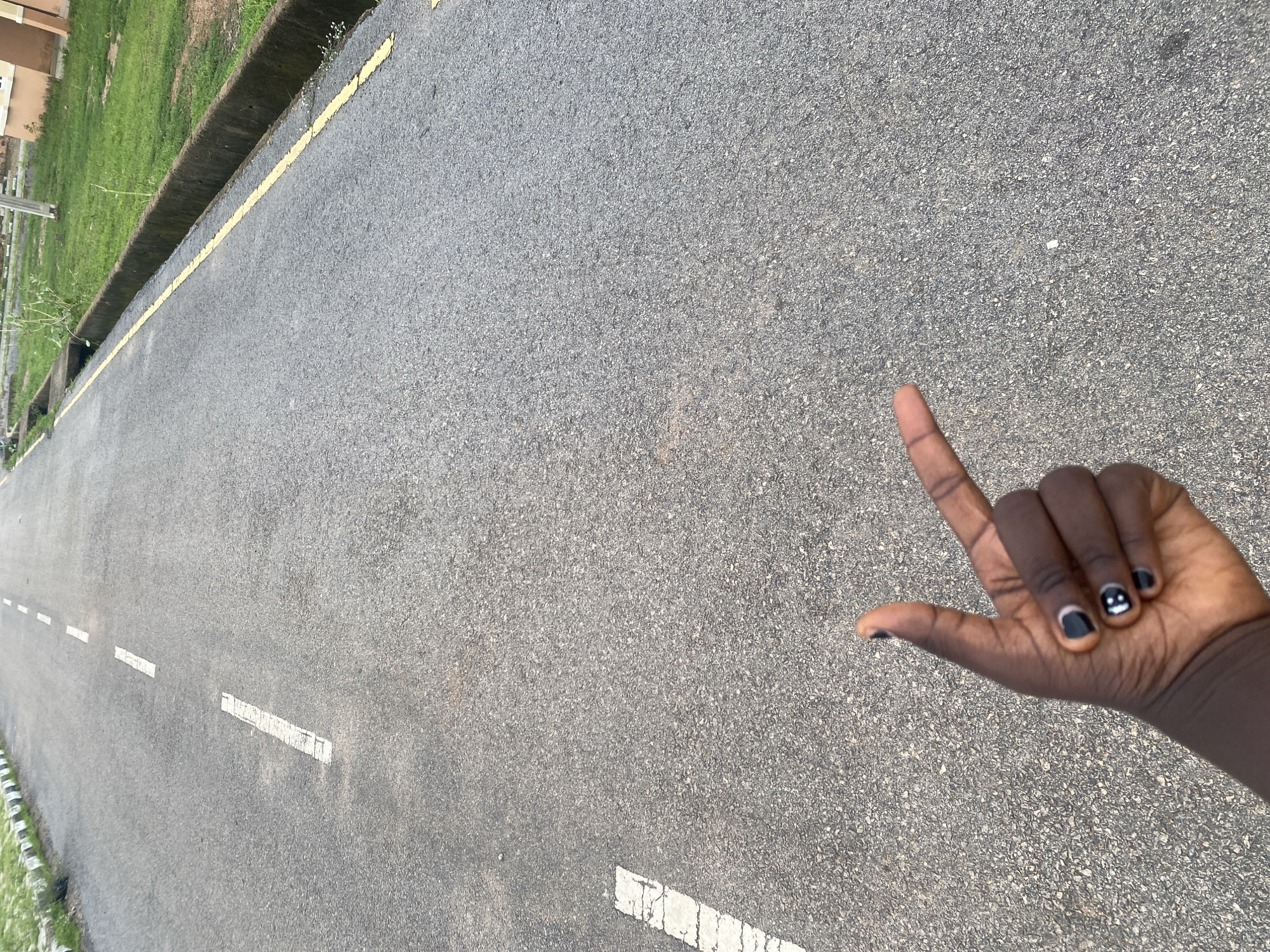 A low-angle outdoor shot depicts a person's left hand with manicured black fingernails, some adorned with white smiley faces, pointing towards the right of the frame. The hand is positioned over an asphalt road. White dashed lines are visible on the road, indicating lanes. To the right of the road, a yellow line demarcates a concrete drainage channel, beyond which is a grassy area. The scene appears to be located on a road within Bulamare, Nigeria.