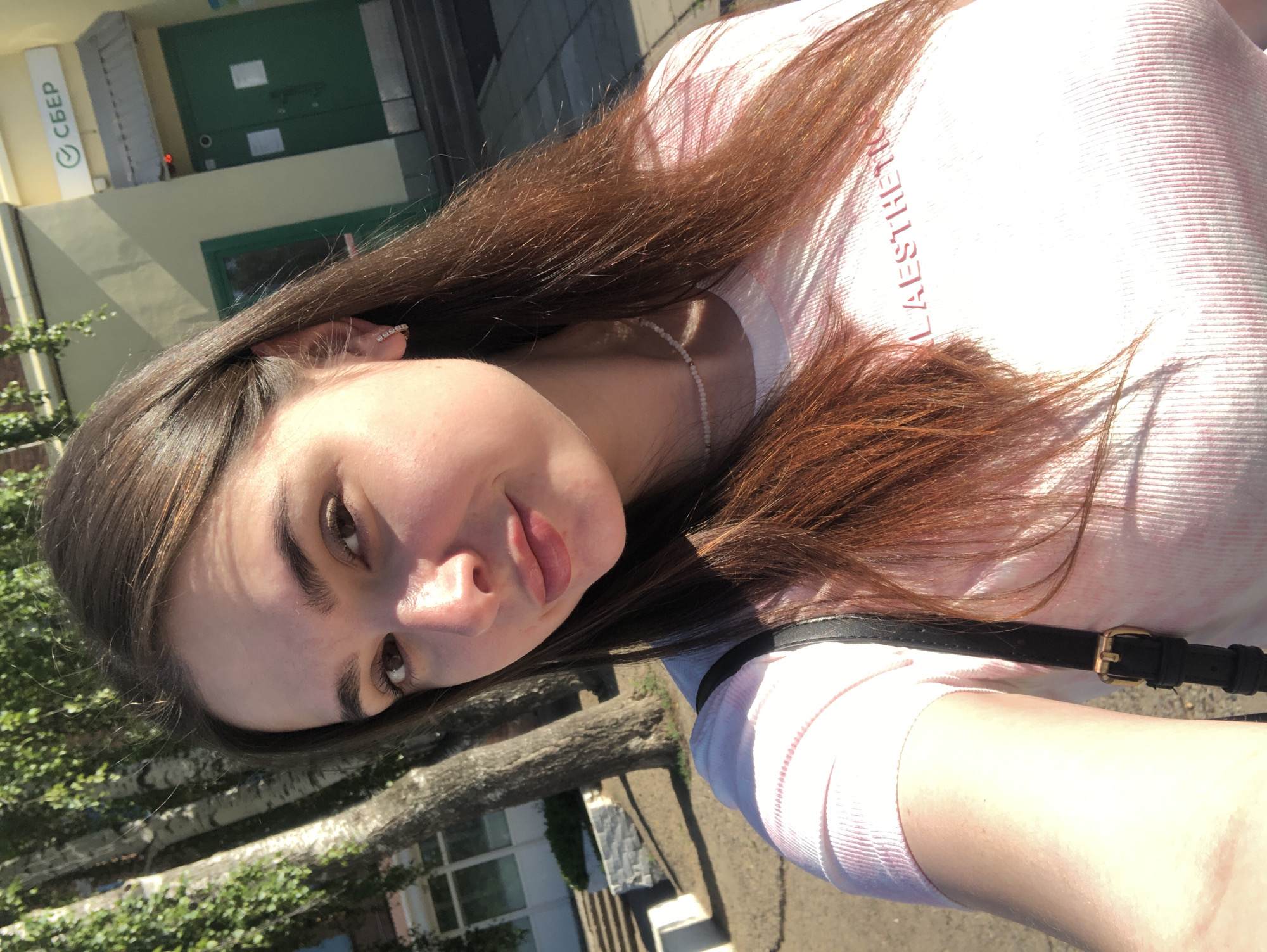 A young woman is taking a selfie outdoors, with a building and trees in the background. She has long, brown hair and is wearing a pink t-shirt and a thin necklace. Her right hand is visible, and she is carrying a black purse with a gold buckle. In the background, a building with a yellow facade and green doors is visible, with a sign that reads "СБЕР". This appears to be a Sberbank branch, a common sight in Russian cities. The lighting suggests it is daytime, with sunlight casting shadows. The presence of the Sberbank sign strongly suggests the location is in Russia, and given the context provided, it could be Noviy.