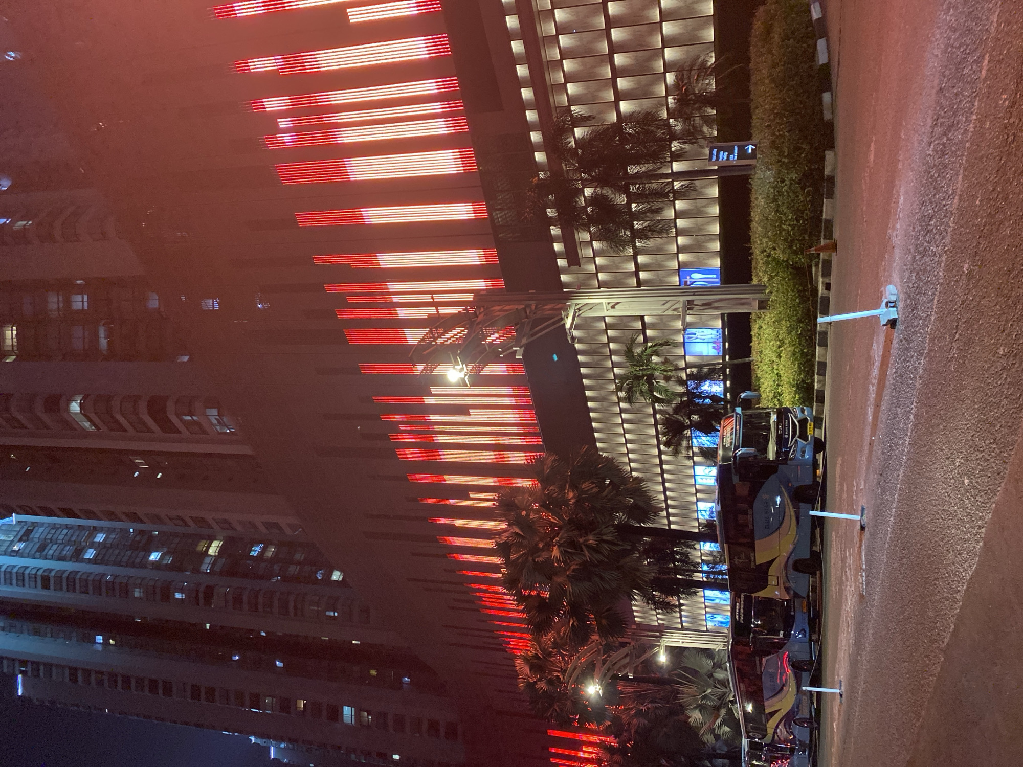 This image shows a nighttime scene in Tangerang, Indonesia, likely at the AEON Mall BSD City. In the foreground, a paved road leads towards several parked blue and yellow buses. The buses have "BLUE STAR" branding. Beyond the buses, there is a well-lit building with rows of illuminated panels and a prominent facade decorated with vertical red and white lights that create a striking visual effect. Palm trees are scattered in front of the building. In the background, tall skyscrapers with illuminated windows dominate the skyline, indicating a dense urban environment. The overall atmosphere is vibrant and modern, illuminated by artificial lights against the dark night sky.