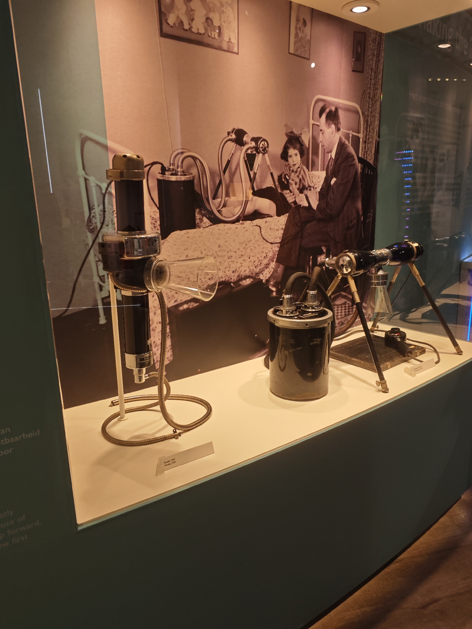 This image shows an exhibit inside a museum. The exhibit features historical medical equipment displayed on a white platform in front of a large photograph of a man and a child in a bedroom setting. The man is seated next to the child, who is sitting on a bed. The photograph appears to be from the early to mid-20th century.
In the foreground, there are two distinct pieces of medical equipment. On the left is a tall, black and silver device with a brass cap and a glass funnel at the top, connected by a flexible metal hose to a smaller black cylinder at its base. On the right, there is another apparatus with a black cylindrical base, and a tripod-like stand supporting a glass tube with a light source inside. This device is also connected to the base via hoses. A small label is visible beneath the equipment on the right, which reads "Revox; 1939; Ronden, 1939."
The background photograph depicts a room with a metal-framed bed, a window with curtains, and framed pictures on the wall. The lighting in the exhibit is warm and focused, highlighting the objects on display. The overall environment suggests an educational display about the history of medicine.
The text on the left side of the image, partially visible, is in Dutch and discusses "vatbaarheid" (susceptibility) and "vooruit" (forward). The text on the upper right, also in Dutch, is cut off but appears to be part of a title related to "making the in" and "Het onzichtbare zichtbaar maken" (making the invisible visible). This likely refers to the historical context of the exhibited items.
While the image itself does not explicitly state the location, a search of museum exhibits related to historical medical equipment and the phrase "Het onzichtbare zichtbaar maken" points to the Philips Museum in Eindhoven, Netherlands. The museum's focus is on the history of Philips, a company that has been involved in medical technology.