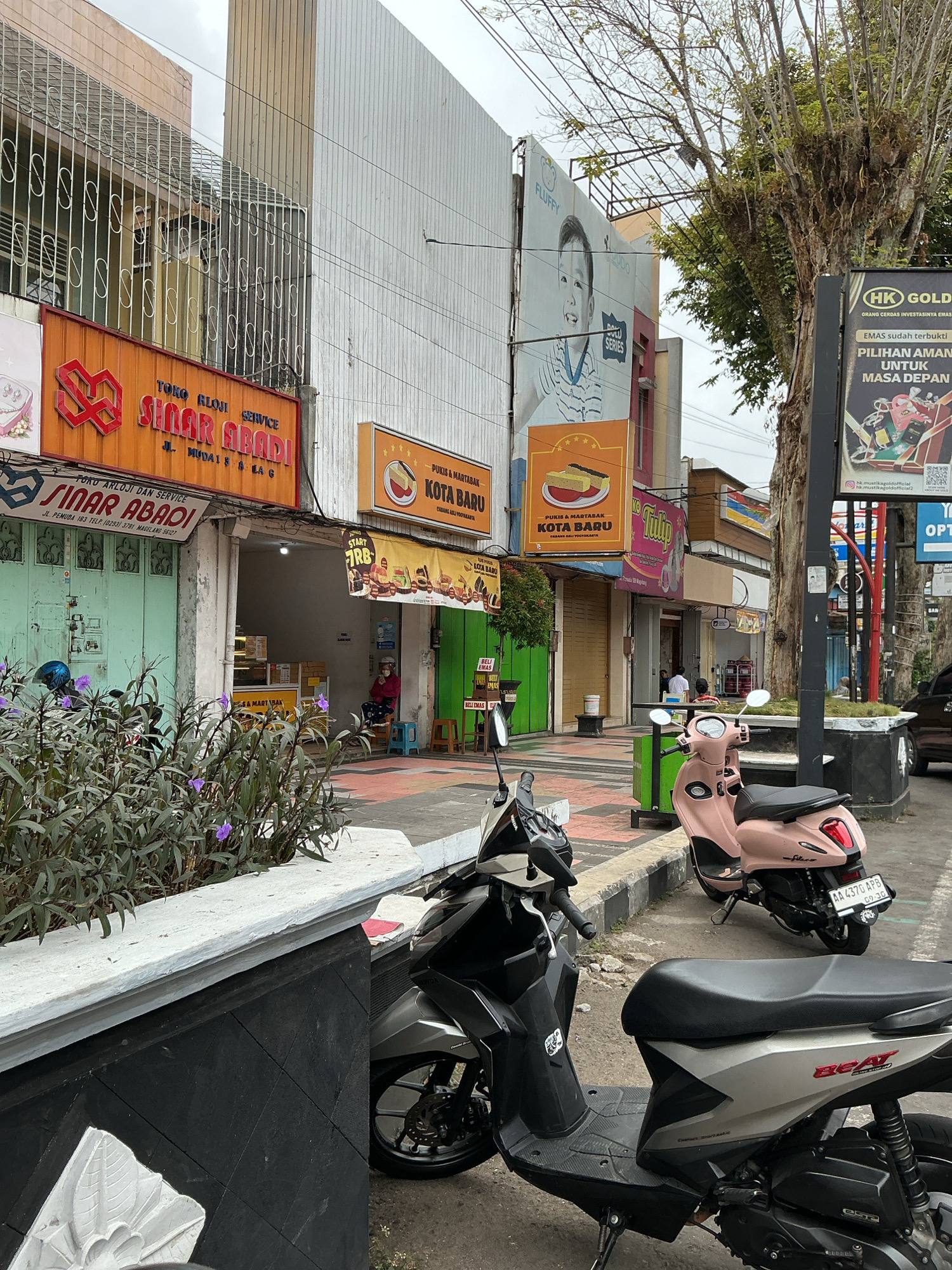 The image depicts a street scene in Magelang, Indonesia, featuring several businesses lining a sidewalk. In the foreground, two scooters are parked, one a dark gray Honda Beat and the other a light pink scooter. The sidewalk is paved with patterned tiles, and a low, white-topped wall with a black base runs along the edge. Beyond the wall, there are plants with purple flowers.
Several shops are visible. The prominent orange sign on the left reads "SINAR ABADI" with the text "TOKO RLOJI SERVICE" above it and an address "JL. MUDAI & KILAG" below. To its right, a shop with a green facade also displays "SINAR ABADI" with a phone number and address. Further down the street, another shop has an orange sign with "KOTA BARU" and "PUKIS & MARTABAK" printed on it. A large billboard above these shops features a picture of a smiling child and the text "FLUFFY". To the right, a tall tree with bare branches stands next to a sign for "HK GOLDEN".
A person is seated on a blue stool outside one of the shops, facing away from the camera and towards the interior of the store. Other individuals are visible inside the shop. Electrical wires are strung overhead across the scene, and the sky is overcast. The environment appears to be a commercial area with a mix of shops and pedestrian traffic.