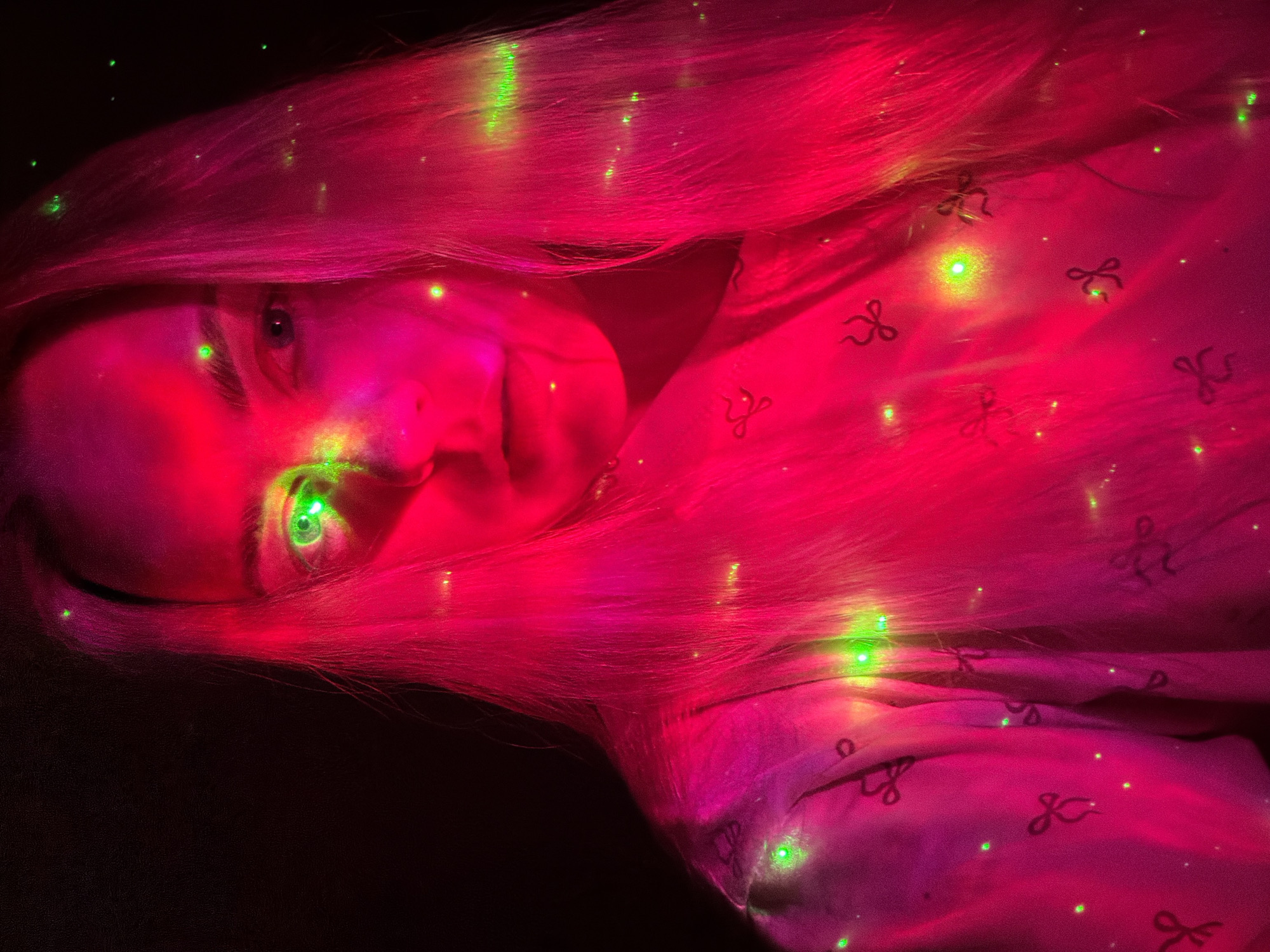 An eye-level portrait captures a person, likely a woman, in an indoor setting illuminated by vibrant artificial lighting. The scene is dominated by intense red and magenta hues, with numerous small, bright green laser-like dots scattered across the subject's face, hair, and clothing.
The person's face is positioned on the left side of the frame, looking towards the viewer with a neutral expression. Long, flowing hair, appearing bright pink or magenta under the lights, cascades across the image. A notable detail is a distinct green laser dot directly on the iris of the subject's visible eye, creating a striking, glowing effect. Additional green dots are visible on the skin and hair.
The subject is wearing a light-colored garment, possibly pajamas or a shirt, which features a pattern of small, dark bow-tie shapes. This clothing is also bathed in the red light and illuminated by several green dots. The background is dark, suggesting a low-light environment where the artificial colors and points of light create a surreal and captivating atmosphere. No text is visible in the image.