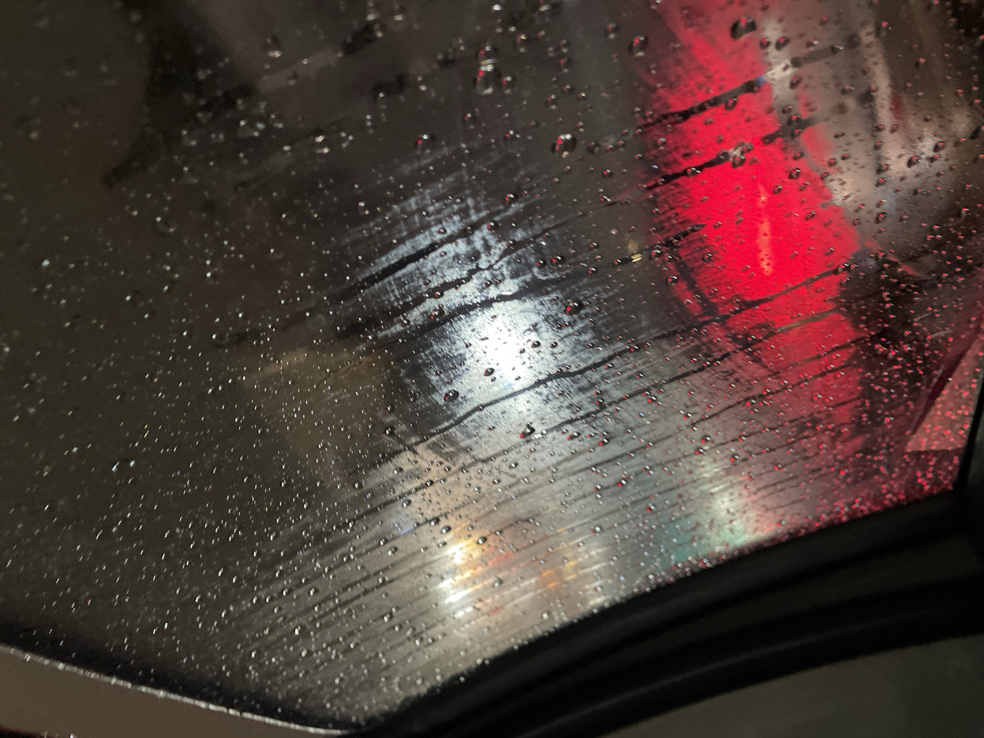 The image shows a view through a wet sunroof, likely of a car, at night. Raindrops cover the glass surface, obscuring the outside scene. Red and white lights are visible through the rain, suggesting a city environment. Given the request to locate this in Buenos Aires, Argentina, these lights could be from traffic signals, streetlights, or building illumination. No people are visible, the focus is on the rain-obscured cityscape through the glass. The overall environment seems urban and wet.