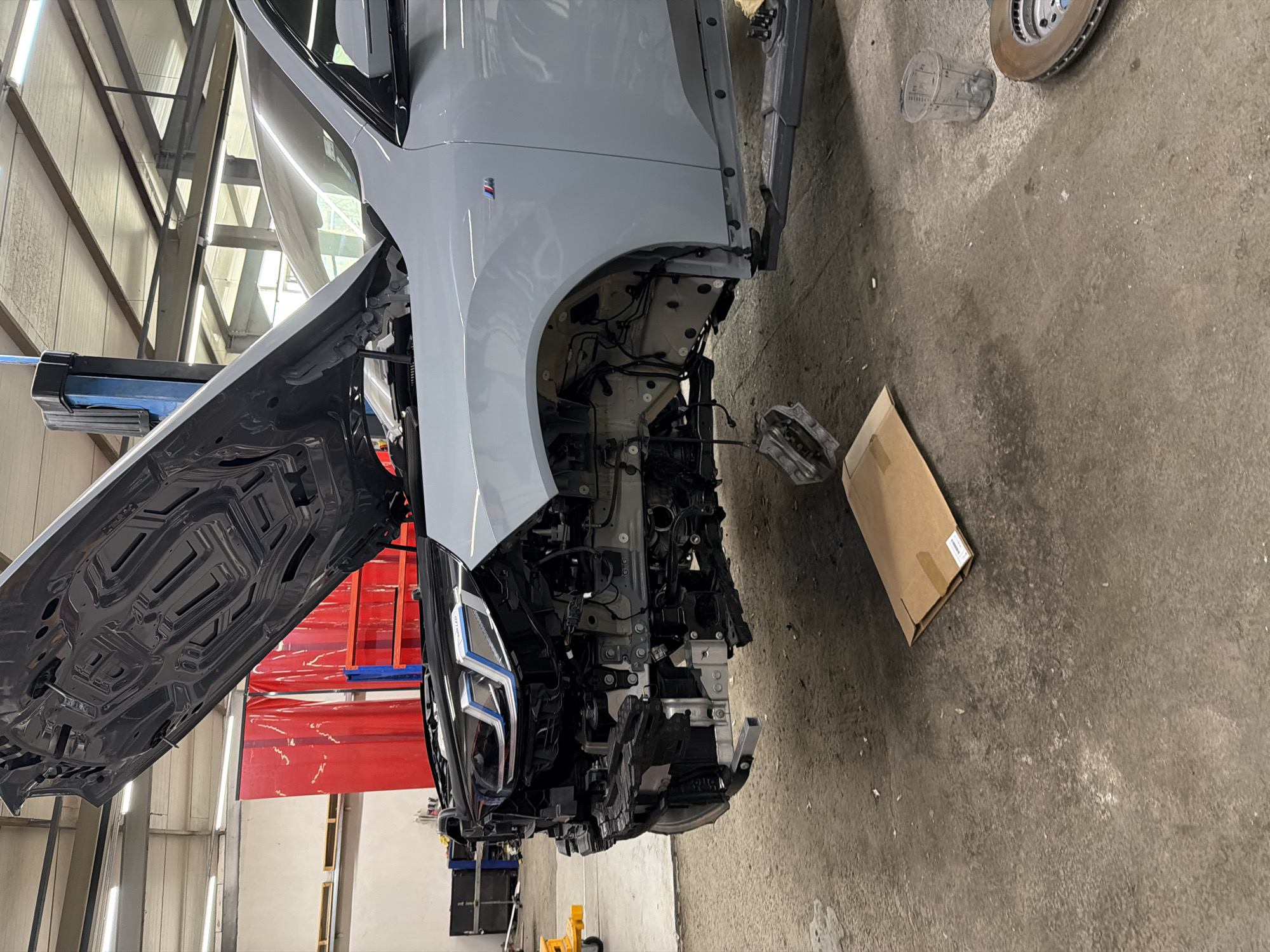 An overhead shot captures a light grey car, identified by a BMW M-series badge on its front fender, in a state of significant disassembly within an automotive workshop. The vehicle's hood is fully open and raised, revealing its dark, structured underside. The entire front bumper and fender assembly have been removed, exposing the internal frame, complex wiring harnesses, and various mechanical and electronic components. The car's headlights, featuring distinctive blue accents, remain attached to the exposed front structure.
On the concrete floor to the right of the car, a rusted brake rotor and a silver brake caliper are visible, suggesting components that have been removed or are awaiting installation. A clear plastic measuring cup and a brown cardboard box are also on the floor. The background reveals typical workshop elements: high ceilings with industrial metal beams, bright fluorescent lighting, and a partial view of a red fabric screen or partition. A black and blue arm of a car lift is visible supporting the vehicle on the left. The scene depicts an ongoing repair or maintenance operation. No individuals are present in the image.