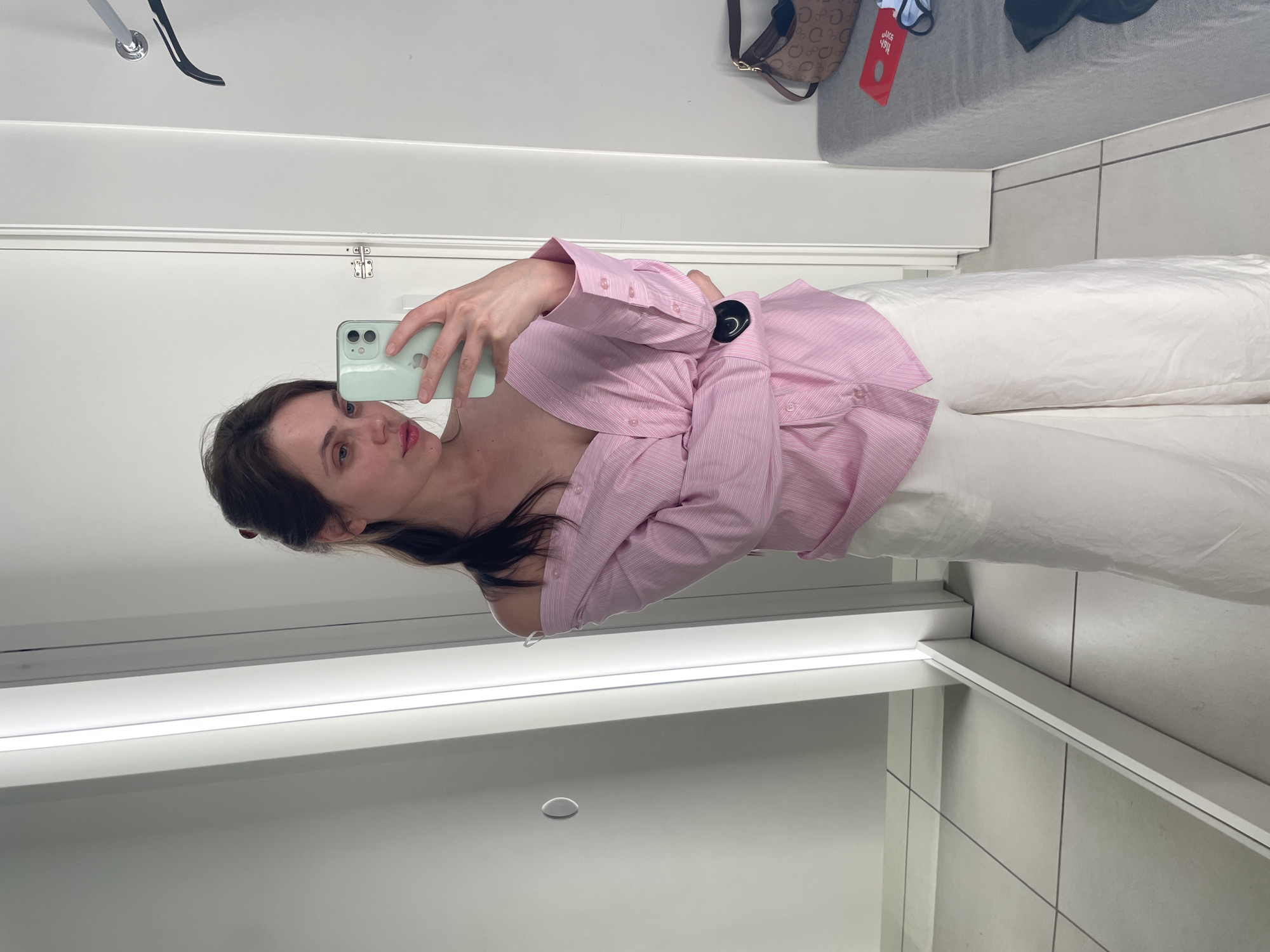 A woman is taking a selfie in a well-lit fitting room. She is wearing an off-the-shoulder pink and white striped button-down shirt and white wide-leg pants. She has her hair pulled back in a ponytail and is holding a mint green smartphone in her right hand. The fitting room has a full-length mirror on the left, a white door on the right, and a grey bench with a brown patterned bag and a red tag on it. The floor is tiled in a light color. The scene appears to be inside a retail store.