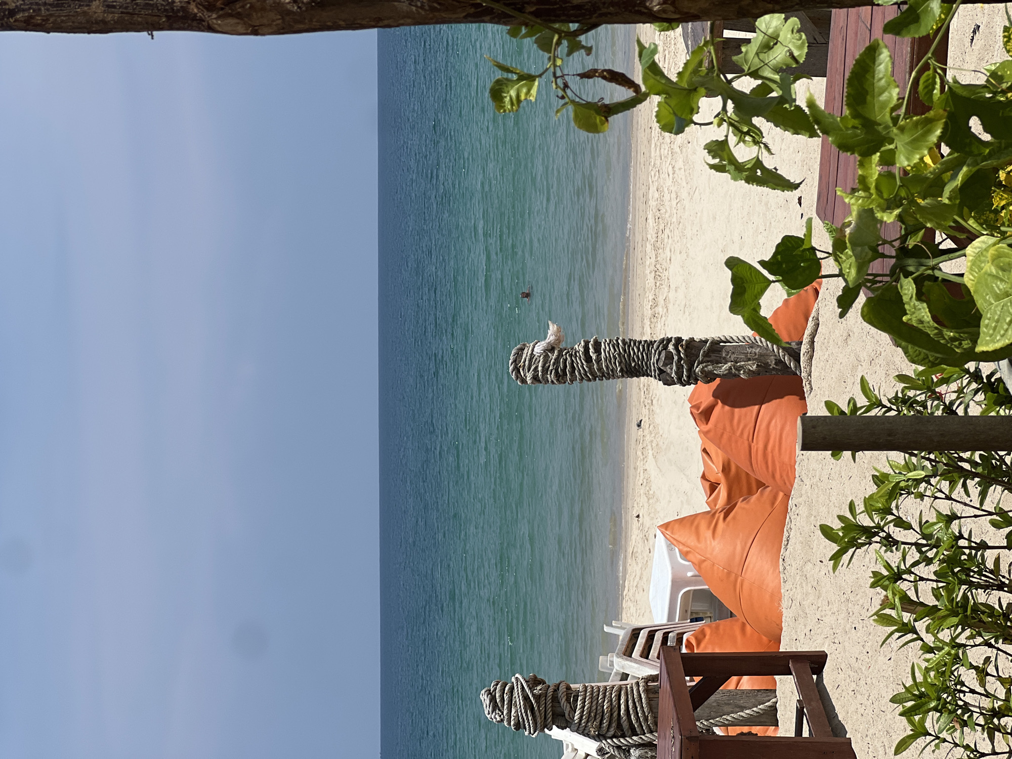 The image depicts a serene beach scene, viewed from a slightly elevated position, possibly a deck or balcony, with elements of a beachside establishment in the foreground. The perspective is framed by a rustic wooden beam at the top and lush green foliage on the right side.
In the immediate foreground, to the right, several orange beanbag chairs and white plastic chairs are arranged on a light sandy beach, suggesting a leisure or relaxation area. Parts of a wooden structure, possibly a railing or post, wrapped extensively with thick rope, are prominent. A small, light-colored bird is perched on one of these rope-wrapped posts, facing the sea.
The midground features a wide expanse of light, finely textured sand, marking the coastline. Beyond the beach, the calm, clear turquoise-blue sea stretches towards the horizon. A lone person, appearing as a small dark figure, is swimming in the sea, quite a distance from the shore. The sea gradually transitions into a pale blue, cloudless sky that occupies the upper left portion of the image.
The weather appears sunny and pleasant, with bright natural light illuminating the scene. The time of day is likely late morning or early afternoon, given the brightness. There is no visible text in the image. The overall impression is one of a tranquil tropical or warm-climate beach getaway.
