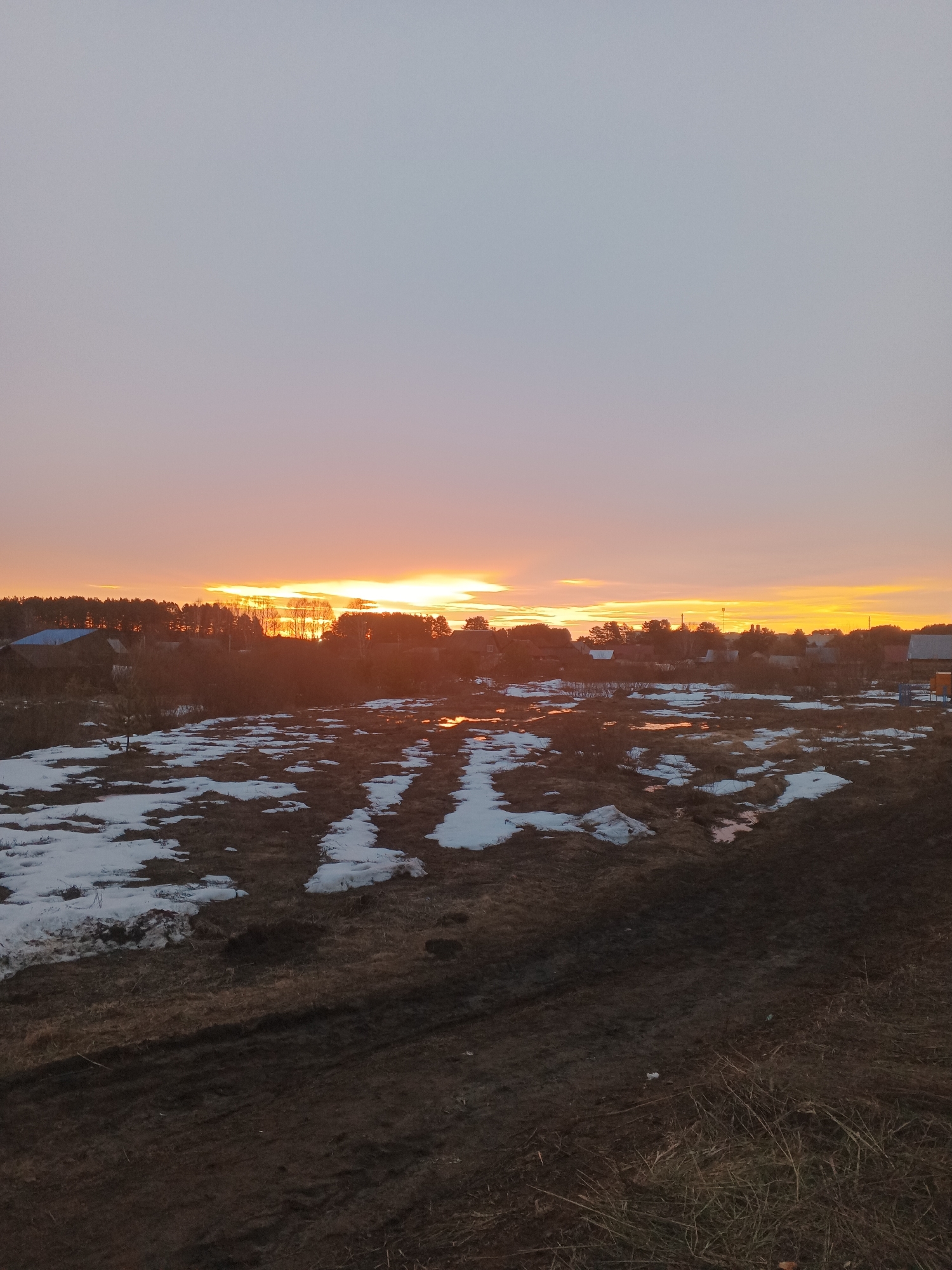 The image depicts a rural landscape at sunset, possibly in or near Rusalevka, Russia. The sky transitions from a pale blue-gray at the top to orange near the horizon. The setting sun casts a bright glow behind a line of trees and rooftops, suggesting a small village or settlement. The foreground is a field with patches of melting snow and standing water. The ground appears muddy and there are visible tire tracks. No people are visible in the scene.