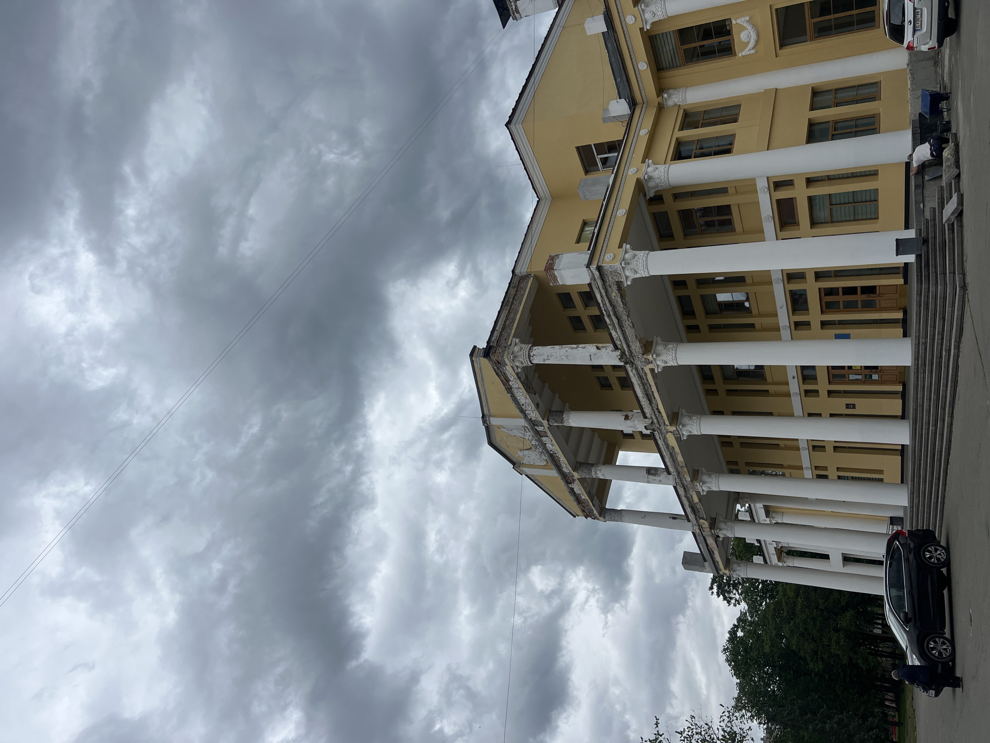 The image captures a large, classical-style building in Kyiv, Ukraine, under a heavily overcast sky. The building, painted pale yellow, features a prominent portico supported by multiple white columns with ornate capitals. Signs of wear are visible, with paint peeling from the columns and cornices. Numerous rectangular windows with dark frames are present across the facade.
The sky is dominated by thick, dark gray clouds, creating a gloomy, dull daytime atmosphere. Two thin power lines stretch diagonally across the upper portion of the frame. In the foreground, a paved area leads up to the building's entrance via a set of stairs.
A dark-colored SUV or hatchback is parked near the building on the right side of the image. A person wearing a light-colored top is partially visible beside the car, appearing to be bending over. Green trees are visible in the background, behind the vehicle. The car bears a Ukrainian license plate, partially visible as 'AE 5849 HO'.