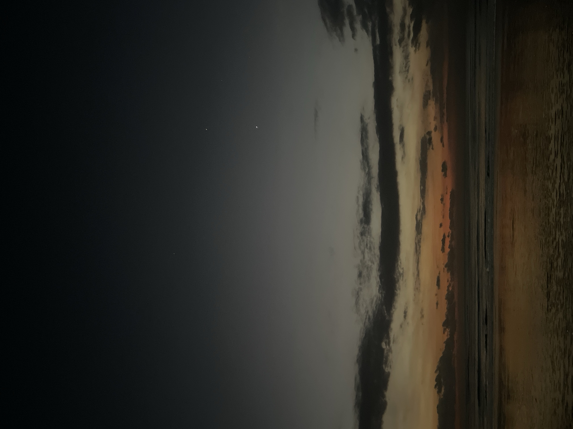 The image, originally oriented vertically but depicting a horizontal landscape, captures a serene night or very early morning scene over a large body of water.
The upper portion of the frame is a dark, almost black sky, transitioning into a lighter grey area further down. Several distinct, bright points of light, likely stars or planets, are visible in the darker sky, with one appearing particularly luminous. Below this, a band of stratified clouds stretches across the middle of the sky. These clouds are predominantly dark grey and black, but some patches near the horizon are illuminated with subtle warm hues of orange and gold, suggesting either a distant light source, a moonrise/set, or very early pre-dawn light.
A clear horizon line separates the sky and clouds from the water below. The lower half of the image features a calm body of water, likely an ocean or a large lake. The water's surface reflects the subtle light from the clouds and sky, appearing in muted dark tones. Gentle ripples or small waves are discernible on the water, adding texture to the otherwise smooth surface.
No people, man-made objects, or specific geographical landmarks are visible. The scene conveys a sense of quiet vastness and the beauty of the night sky over water. There is no visible text.