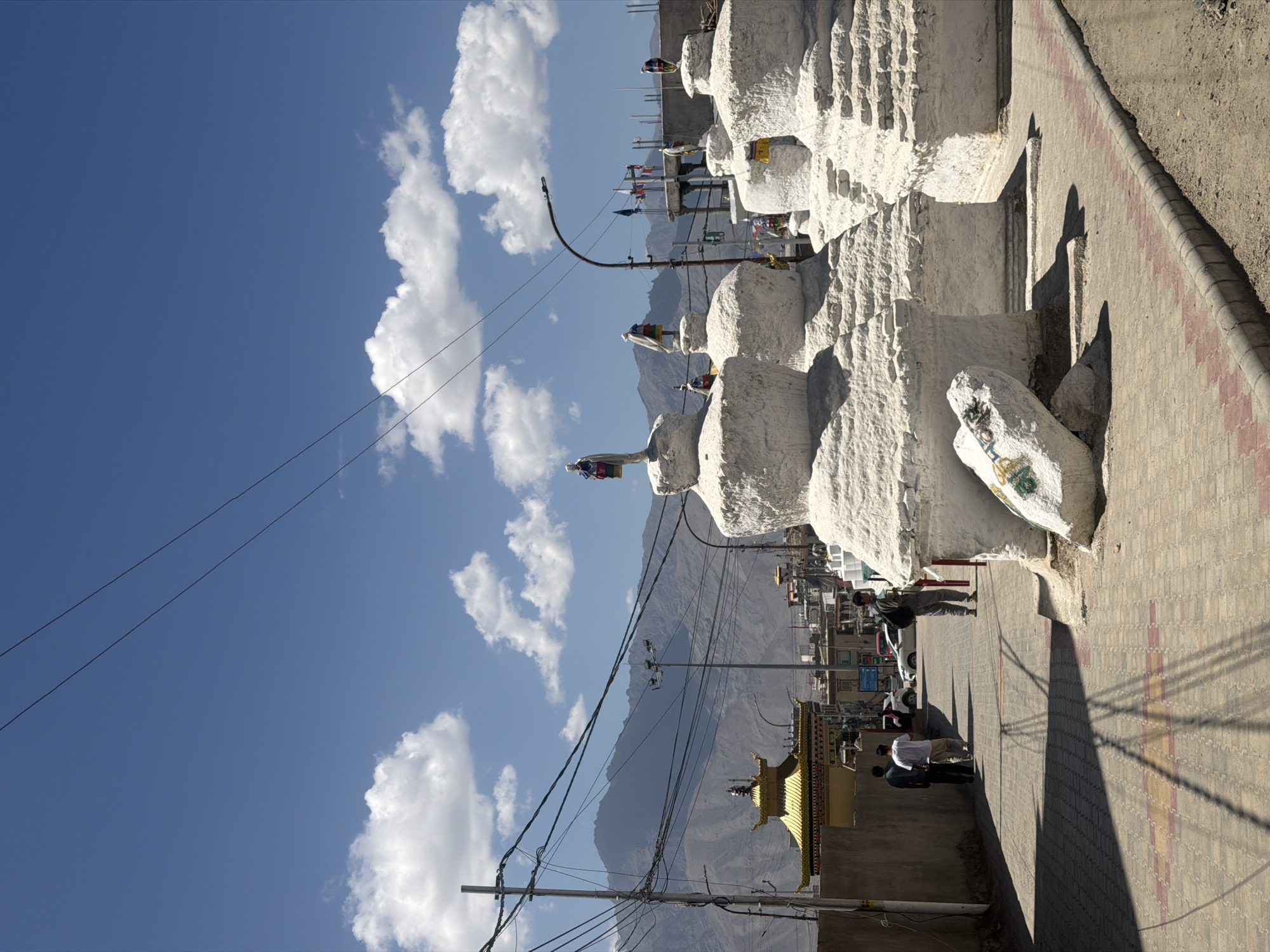 A sunlit, street-level view shows a pathway in Leh, India, leading towards a golden-roofed temple building. In the foreground and right side of the image, several white, stupa-like structures are arranged in a cluster. One of these structures has a large, light-colored rock placed in front of it, with some faint, green painted markings. A paved walkway with alternating red and yellow brick patterns runs along the bottom of the image, bordering a dirt area on the right. Three individuals are walking away from the viewer down the pathway, towards the temple. One individual, in a dark shirt, is followed by two others in lighter colored shirts. Further down the pathway, another individual is walking towards the camera, carrying a backpack. In the background, the golden-roofed temple is visible, along with other buildings and a mountain range stretching across the horizon under a clear blue sky with scattered white clouds. Electrical wires and poles are prominent throughout the scene, indicating urban infrastructure. The overall lighting suggests it is daytime, likely mid-morning or afternoon, with strong shadows cast by the stupa structures and electrical poles.