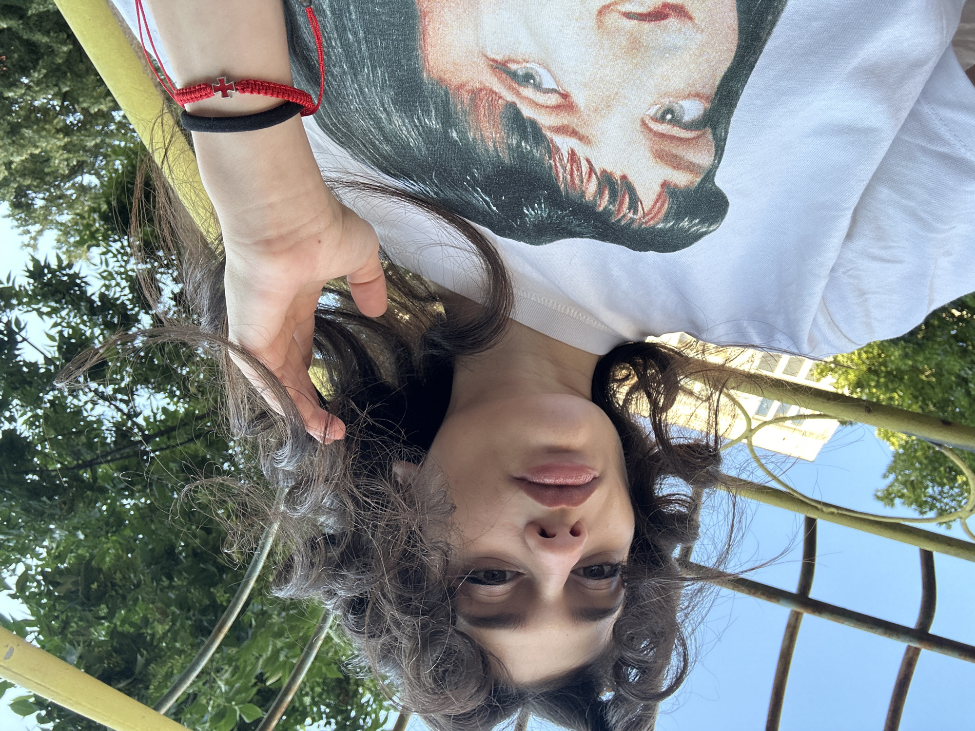 The image shows a young woman with dark, curly hair, looking at the camera. She is wearing a white t-shirt with a graphic of a woman's face on it. A hand with a red bracelet and a black band is visible near her face. The background includes greenery and a yellow structure, possibly part of an outdoor play area or park. The scene suggests an outdoor setting in an urban environment, like a park in Sofia, Bulgaria.