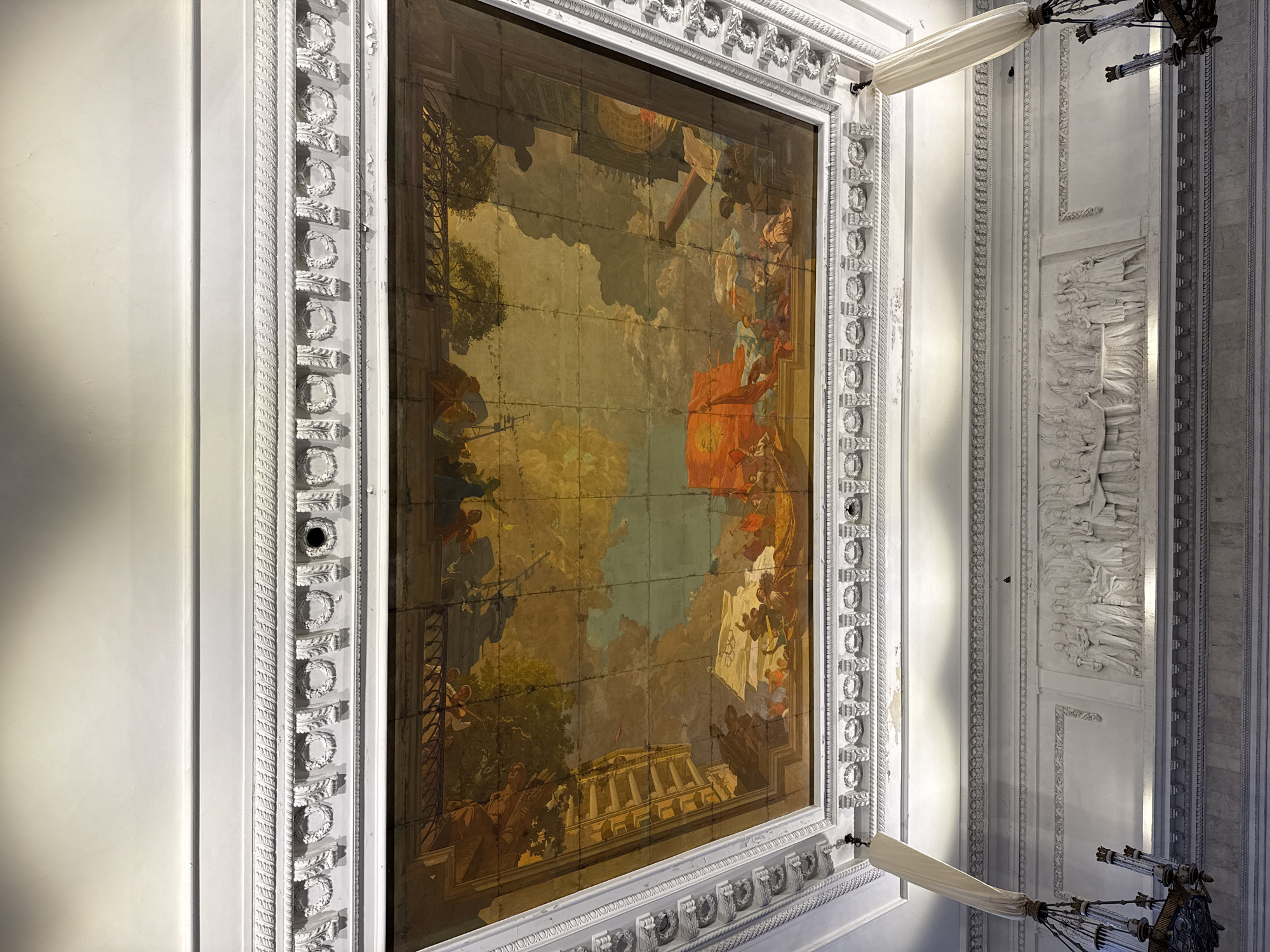 The image shows a grand interior space, likely a museum or historical building, with an ornate frescoed ceiling. The fresco depicts a scene of figures and architecture, possibly allegorical or historical, with vibrant colors of orange, yellow, blue, and green. The ceiling is framed by detailed white plasterwork, including moldings and friezes with laurel wreath motifs. Below the ceiling, on the wall, is a large white bas-relief sculpture featuring a group of figures in classical attire, engaged in what appears to be a formal gathering or ceremony. Two elaborate chandeliers hang from the ceiling, one partially obscured by a white fabric drape. The overall impression is one of opulence and historical significance. This scene is likely located in the State Russian Museum in Saint Petersburg, Russia, given the context of historical art and architecture often found there.