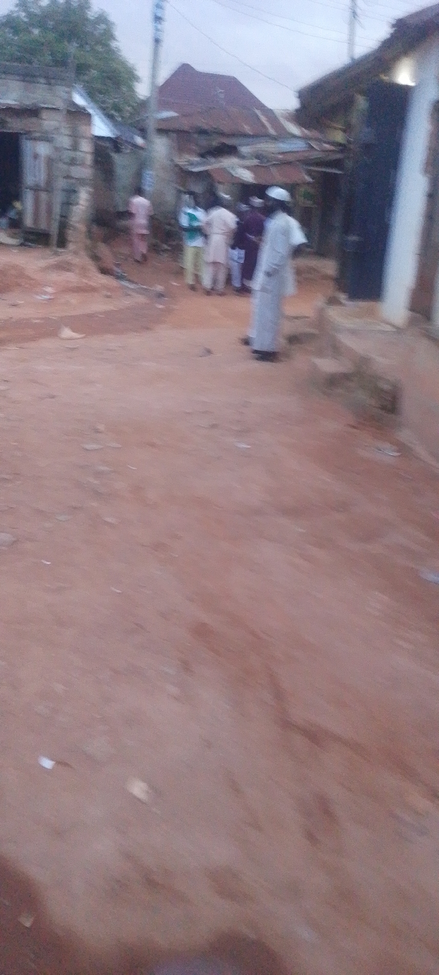The image shows a dusty, unpaved street in Gura Top, Nigeria, with several people gathered in the background. The buildings on either side of the street appear to be made of various materials, including corrugated metal and what looks like brick or concrete. In the foreground, the ground is a reddish-brown dirt, with some scattered debris. A group of people, dressed in traditional Nigerian attire, are standing together in the mid-ground, facing towards the right of the frame. Some of them are wearing white caps. The overall impression is of a bustling, everyday scene in a Nigerian town.