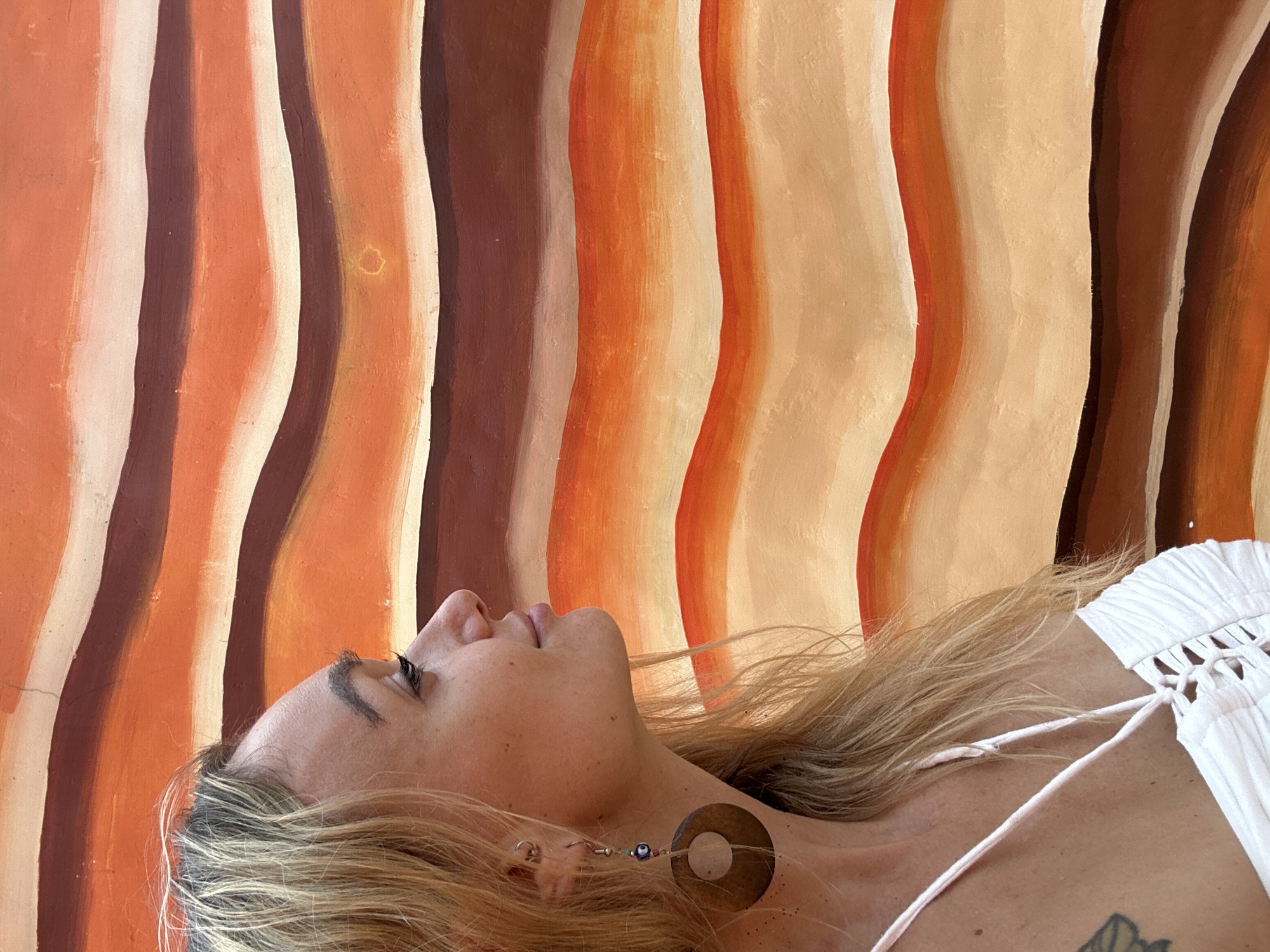 A medium-shot captures a woman lying down with her head tilted back. The background features a vertically-oriented, slightly out-of-focus wall with painted stripes in earth tones of orange, brown, and beige. The woman has light skin, blonde hair, and is wearing a white top with thin straps. An earring is visible on her left ear, and a large, round, wooden earring hangs from it. A tattoo can be seen on her upper chest. The photograph appears to be taken in Mũi Né, Vietnam.