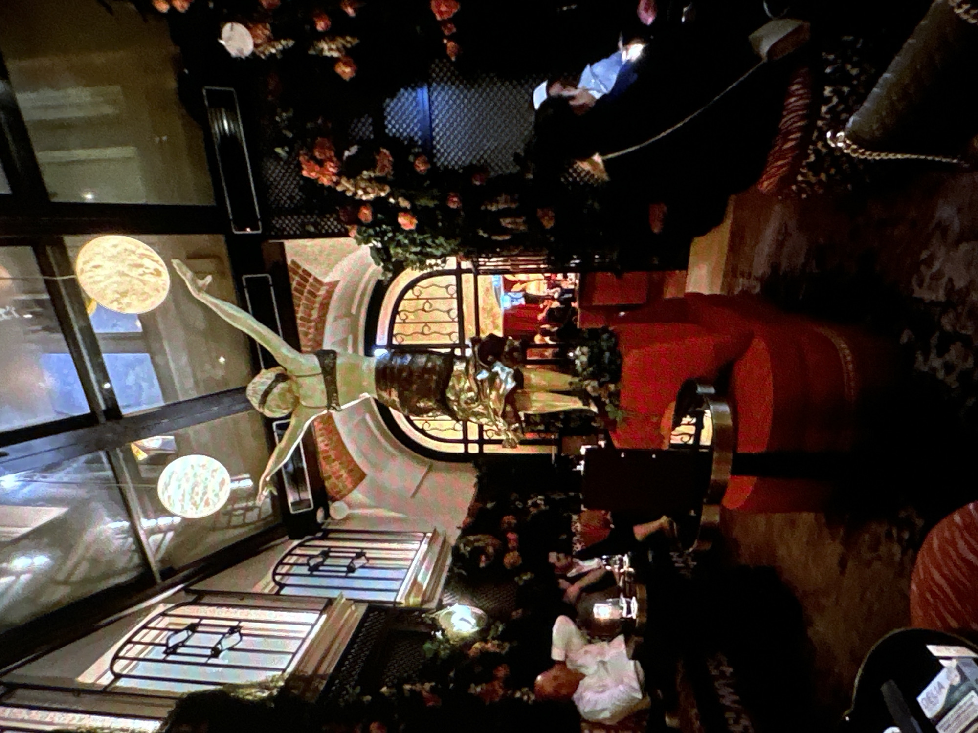 The image depicts the interior of a venue in Chișinău, Moldova, specifically a restaurant named Pegas Terrace & Restaurant. The restaurant features a terrace with decor including artificial flowers and greenery. There are tables occupied by people, some dining and conversing. A gold-colored statue of a woman with arms raised is a central decorative element. The interior design includes architectural details like arched windows and decorative metalwork. Round pendant lights provide illumination.