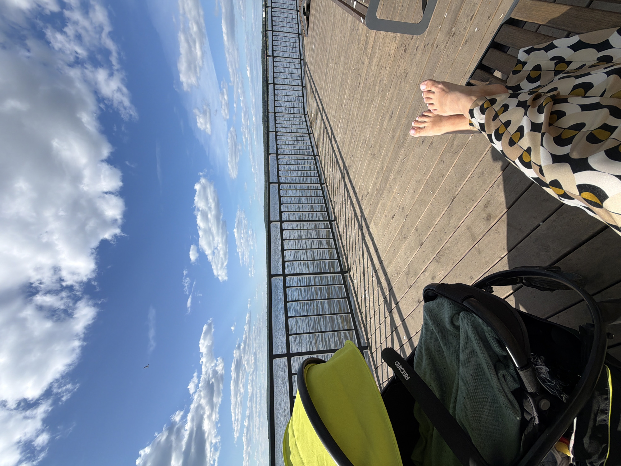 The image depicts an outdoor scene from a wooden deck overlooking a large body of water in Votkinsk, Russia. The upper portion of the frame is dominated by a clear blue sky interspersed with numerous white, fluffy cumulus clouds, with a single small bird in flight against the blue to the left. A dark grey metal railing runs horizontally across the mid-ground, separating the deck from the rippled water, which extends to a distant tree line marking the horizon.
Strong sunlight casts prominent, elongated diagonal shadows of the railing and other objects onto the light brown wooden planks of the deck. To the left, a partially visible baby stroller features a bright neon yellow-green canopy, a dark frame, and the brand name "RECARO" visible on a strap. A dark green, textured blanket is folded within the stroller. To the right, the bare feet and lower legs of an individual are visible, resting upon a dark-framed structure, likely a bench, a portion of which is visible in the lower right. The individual wears wide-leg pants with a distinct abstract geometric pattern in black, white, gold, and brown. The toenails are painted a light pink.