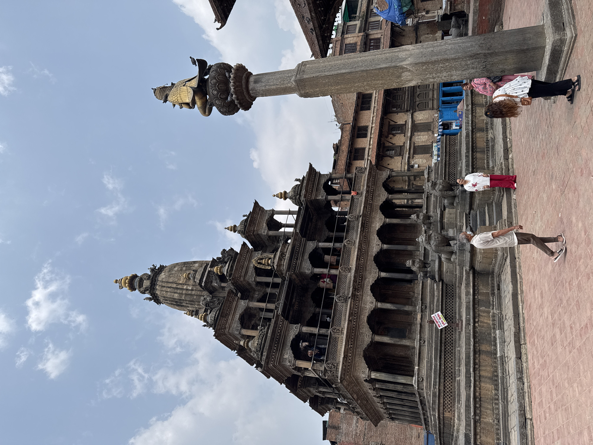 The image shows a vertical view of Patan Durbar Square, a UNESCO World Heritage Site located in Lalitpur, Nepal. The architecture is ornate, featuring multi-tiered pagoda-style buildings constructed with dark wood and brick. Intricate carvings adorn the walls and pillars. A stone column topped with a golden statue stands in the foreground against a cloudy sky. Three people are visible: one man wearing a white shirt and brown pants, and two women wearing white blouses and black pants. They are standing on a brick-paved ground. A white sign is held by the man.