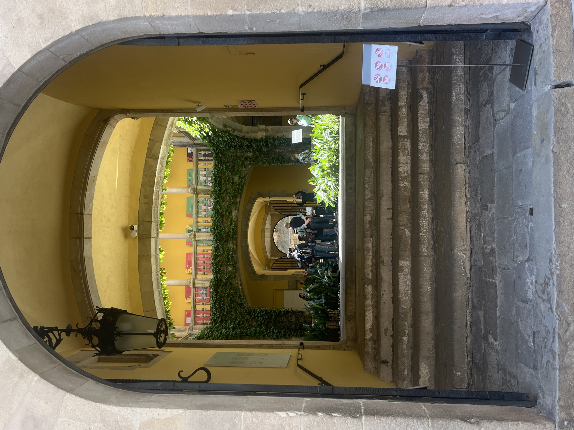 The image captures a vibrant architectural scene, likely a courtyard or cloister, viewed through a stone archway in Barcelona, Spain. The perspective is looking from a relatively darker, stone-arched entrance into a brightly lit, yellow-walled space.
The setting is characterized by a series of receding stone arches and pillars, painted in a warm yellow hue. Lush green ivy cascades down the left wall, adding a natural element to the otherwise stone and stucco structure. On the right, a set of wide, stone steps leads upwards, with a black metal handrail.
In the brightly lit area beyond the initial archway, several people are visible, appearing to be visitors or tourists. They are casually dressed and are gathered in small groups, observing their surroundings. One individual wearing a green and white striped shirt is clearly seen among the foliage.
Notable objects include an ornate, black wrought-iron lantern with a glass shade mounted on the left wall within the foreground arch. High on the left wall, a small security camera is visible. On the upper right, a white square sign displays six red circular symbols, resembling dice faces or a unique logo. Below it, a dark rectangular sign is mounted on the stone wall. On the lower left wall, a white rectangular plaque with illegible text is affixed. Further into the courtyard, along the left wall, several vertical rectangular panels are decorated with red elements and some visible text, specifically the number "5." repeated vertically, suggesting some form of numbering or display. Another arched passage is visible in the distance, further enhancing the depth of the scene.
The scene is bathed in bright daylight, indicating a clear, sunny day, likely late morning or afternoon. The overall impression is one of a historic and well-maintained building, possibly a museum, gallery, or university, attracting visitors.