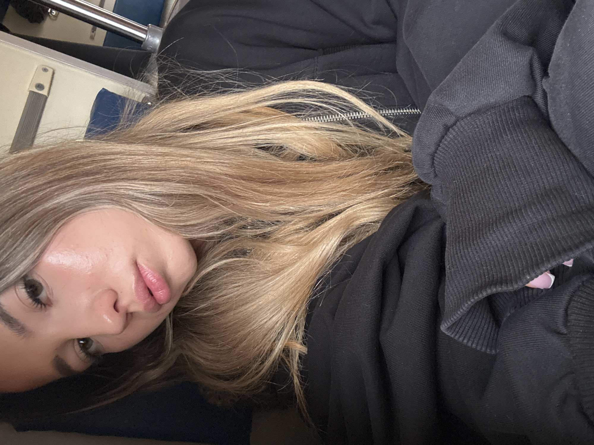 A woman with long blonde hair is lying down, possibly on a train. She is wearing a black hooded jacket. In the background, there are metal poles and a blue seat. The specific location in Moscow, Russia, cannot be determined from the provided image.