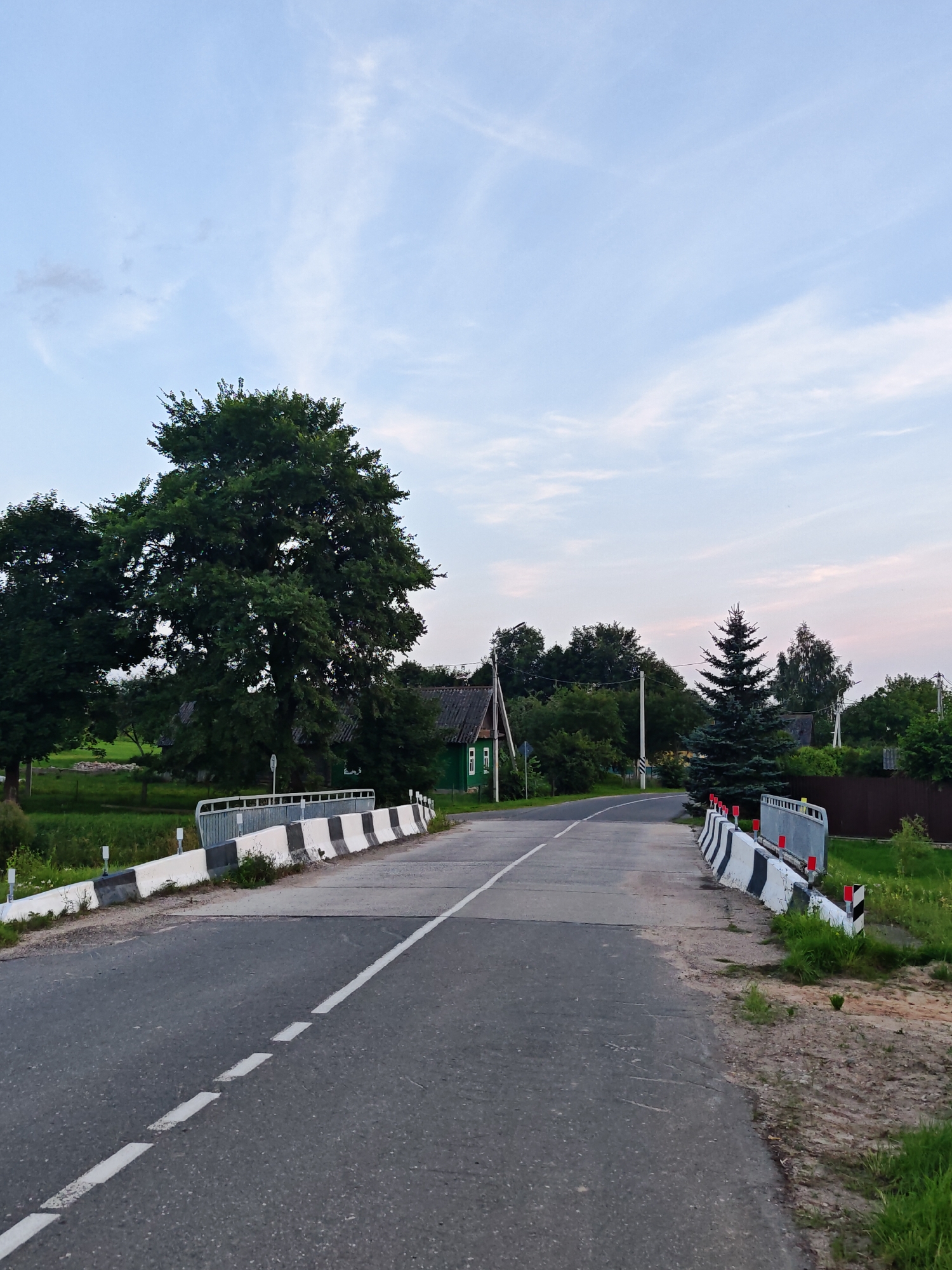 The image depicts a rural road with a bridge in the Валожынскі сельскі Савет, Belarus. The road surface is asphalt with a white dashed line marking the center. On either side of the road, there are concrete barriers with black and white stripes and red reflectors. The bridge railing is visible, with a metal structure and concrete barriers.
To the left of the road, there is a large, mature deciduous tree. Further left, there are fields of green grass and a small structure with a dark roof and green walls, possibly a house or shed, with white trim around the windows.
To the right of the road, a tall, dark green evergreen tree stands prominently. Beyond the evergreen, there are more trees and vegetation, including a brown fence. The sky above is a pale blue with wispy white clouds, suggesting it is either early morning or late evening. The overall impression is a tranquil, natural setting.