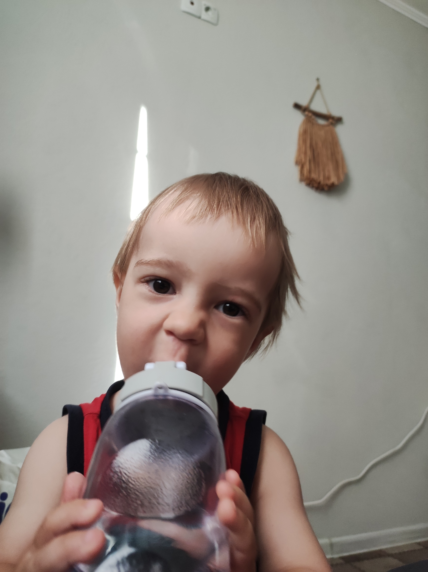 A young child with blond hair is drinking from a clear plastic bottle with a gray lid, making a slightly scrunched-up face. The child is wearing a red shirt with black trim around the armholes. The background features a light gray wall with a bright vertical highlight indicating a window or a light source and a small brown wall decoration that looks like a woven tapestry. A white electrical outlet is visible in the upper left corner. The scene is indoors. The location is Chaikovsky, Russia.