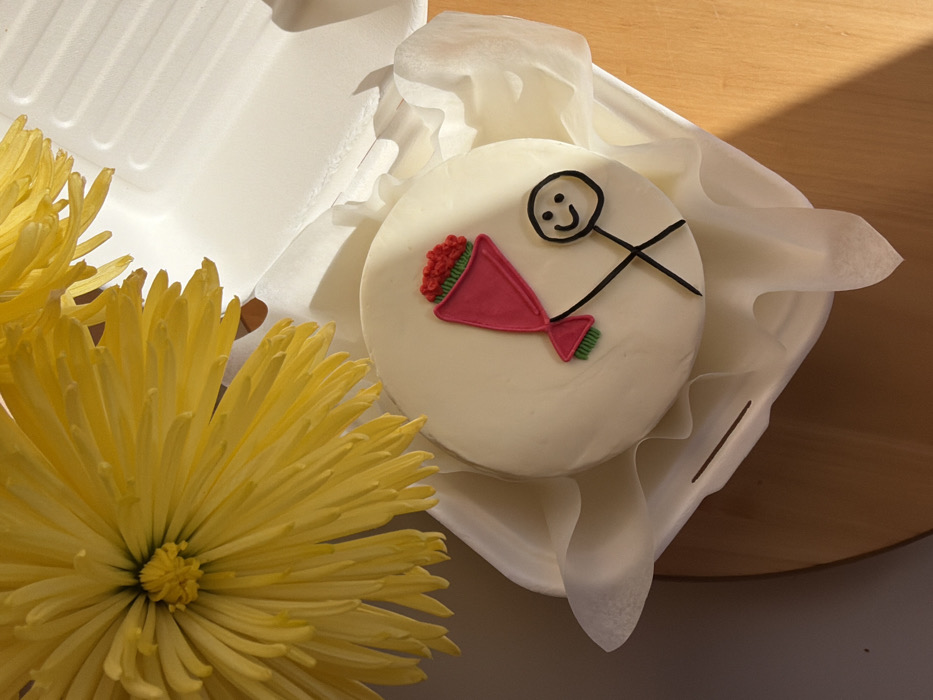 The image displays an overhead view of a small, round cake with white frosting, resting inside an open, white foam takeout container lined with white tissue paper. The cake is decorated with a black frosting stick figure featuring a smiling face, holding a pink-wrapped bouquet of red rosettes and green leaves. A cluster of yellow chrysanthemum flowers occupies the upper left quadrant of the frame, partially obscuring the container, with distinct petals and a visible central disc. The entire arrangement is situated on a light brown wooden surface. Strong, focused lighting casts clear shadows on the cake and container from the right. A solid light grey wall is visible in the background to the left. No individuals are present. The image provides no visual indicators to confirm a specific location within Tel-Aviv, Israel.