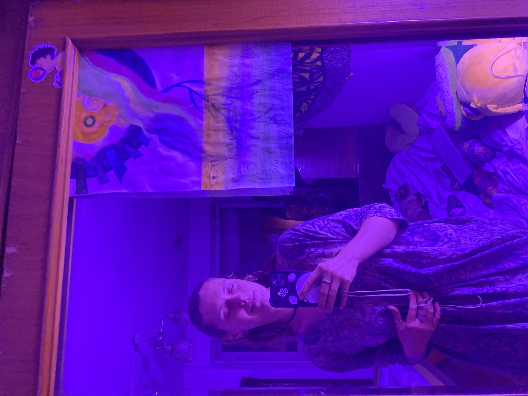A person is taking a selfie in a mirror. The person is wearing a patterned dress and has their eyes closed and a slight smile on their face. They are holding a smartphone in their right hand, and their left hand is resting on their stomach. There is a drawing taped to the mirror above the person's head. The room is lit with purple light. The location is not specified as being in Almaty, Kazakhstan.