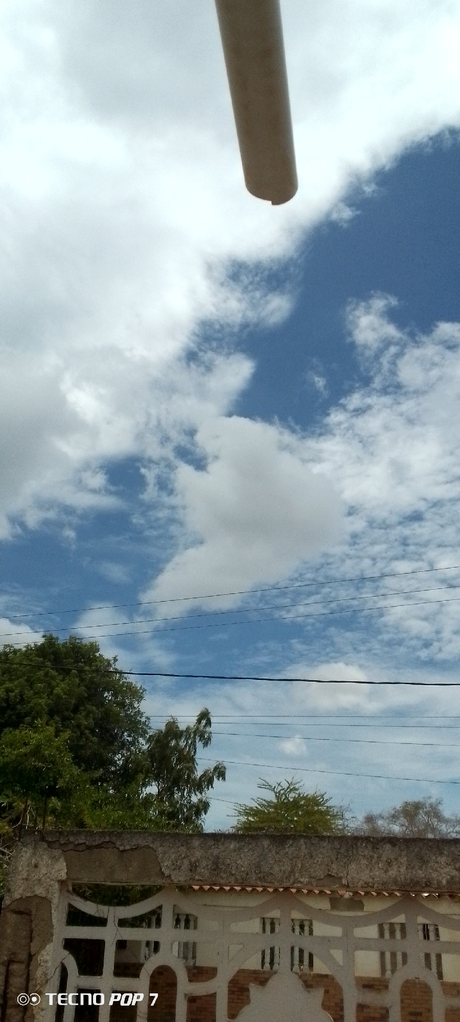 The image depicts a partly cloudy sky over a residential area in Altagracia, Venezuela. A cylindrical object (possibly a pole or pipe) is in the upper center of the frame, obscuring part of the sky. Below the sky, power lines stretch horizontally across the scene. Green trees are visible behind a concrete fence or wall. The fence is white with a decorative pattern. Cracks and damage are visible on the fence's surface. A watermark indicating "TECNO POP 7" is in the lower left corner of the frame.