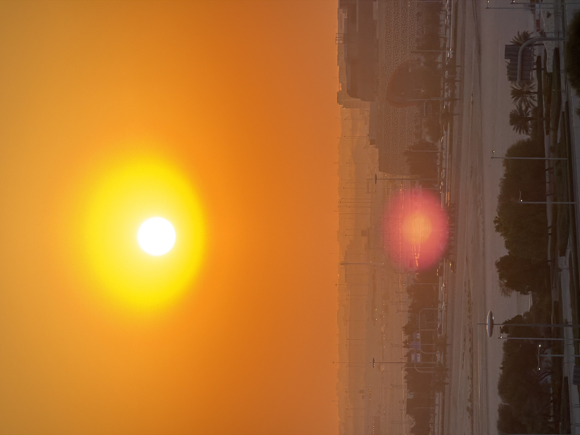 The image depicts a bright, hazy sunset over a city landscape. The sun, a large, glowing orb, dominates the left side of the frame, casting a warm, orange hue across the sky. In the distance, a hazy silhouette of buildings is visible, suggesting an urban environment. Palm trees and streetlights are scattered throughout the scene, indicating a potentially arid or tropical location. The right side of the image shows a road with some cars and a faint lens flare reflecting a reddish glow. The overall atmosphere is serene and bathed in the soft light of dusk. The time of day is clearly sunset due to the sun's position and the warm colors. The weather appears to be hazy, as evidenced by the softened outlines of distant objects and the diffused sunlight. There are no people visible in the image.