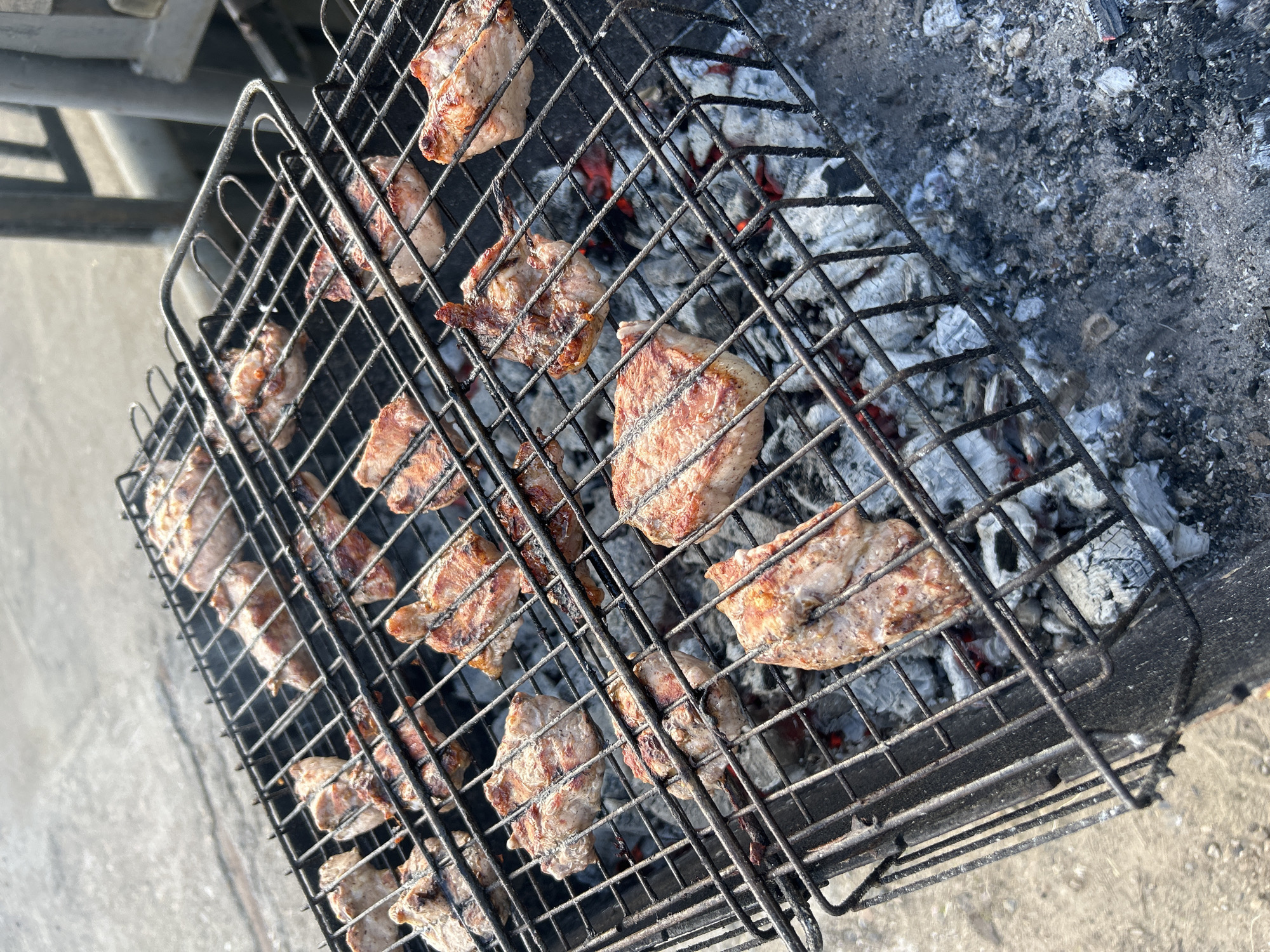 A top-down, slightly high-angle shot depicts a barbecue grill with meat cooking over hot coals and ash. The grill is a double-sided wire rack type, enclosing the meat to facilitate turning. The meat pieces are generally small and appear to be pork, judging by their color and texture. They are arranged on the grill and exhibit varying degrees of cookedness, with some pieces showing more browning and grill marks than others. Beneath the grill, smoldering coals and ash are visible, indicating recent or ongoing use. The background consists of concrete or stone, which is likely part of an outdoor cooking area. The image was taken outdoors, in daylight. The location in the city of Chaikovsky, Russia, cannot be confirmed.