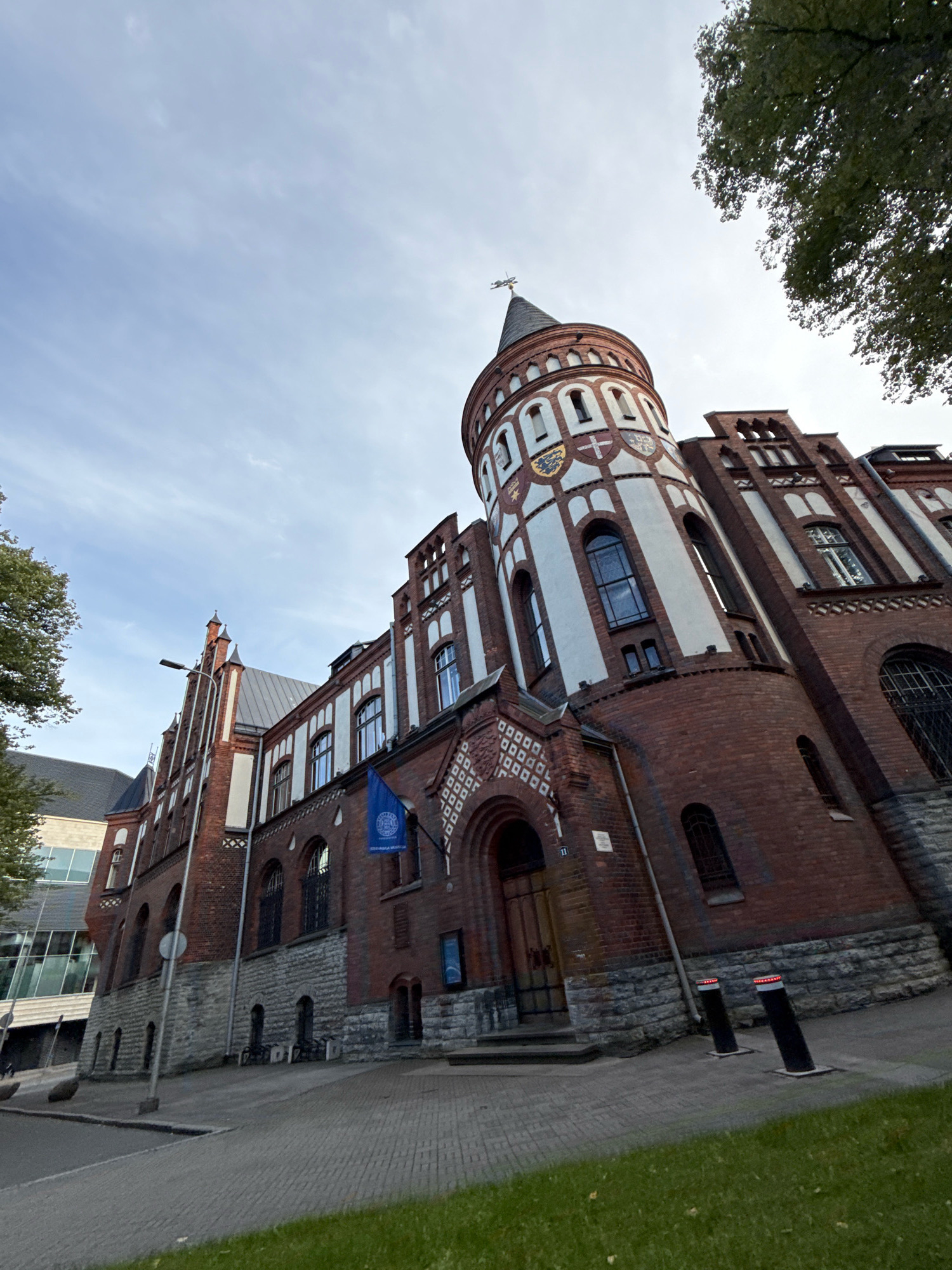The image shows the House of the Blackheads in Tallinn, Estonia. The building is made of red brick and has a distinctive tower with a pointed roof. There is a blue flag hanging on the front of the building. The sky is cloudy. The building is located on a street with a cobblestone sidewalk. There are trees on the left side of the image and on the right side of the image. There is also a modern glass building in the background on the left.