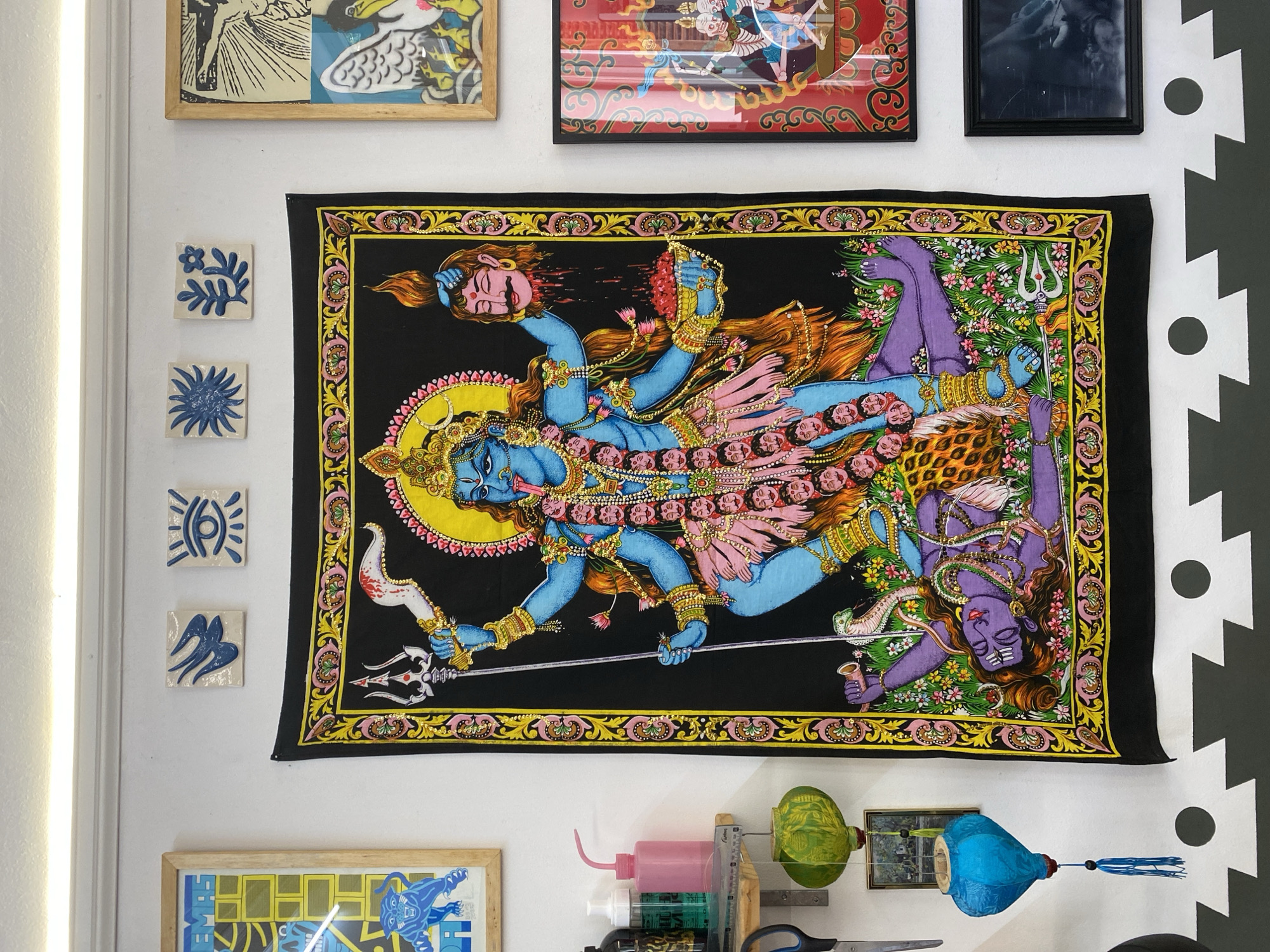 A cluttered interior wall in Buenos Aires, Argentina, displays a variety of items. Three framed pieces hang along the top, with a black-framed photograph on the right. Four small square tiles with blue designs are arranged vertically. A large, rectangular tapestry depicting a multi-armed, blue-skinned Hindu deity occupies the center, adorned with skulls and weapons. Below the tapestry, there's a framed artwork, and an array of items including a pink watering can, spray bottles, a ruler, scissors, and two hanging paper lanterns (one green, one blue). The wall is painted white, with a black and white zig-zag pattern visible on the right side. The overall impression is of an eclectic, bohemian, and artistic space.