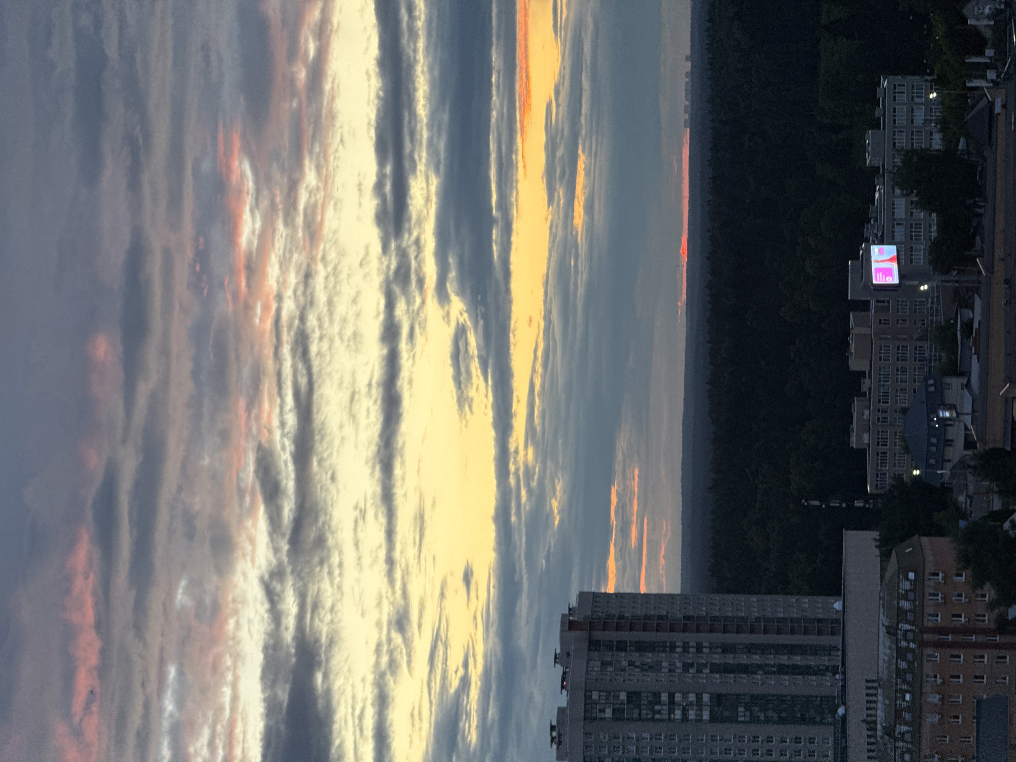 This is a vertical, medium-shot photograph taken from a high vantage point, likely a building, overlooking a cityscape and a forested area. The dominant subject is the sky, filled with dramatic, layered clouds catching the light of what appears to be a sunset or sunrise. The clouds are a mix of dark grays, soft whites, and striking oranges and pinks, suggesting a dynamic interplay of light and shadow.
Below the sky, the scene transitions to a dark silhouette of a dense forest that stretches across the middle ground. On the right side of the frame, the cityscape emerges from the trees. Several multi-story residential buildings are visible, characterized by numerous windows and balconies. The architecture suggests an urban environment.
One of the buildings prominently features a lit billboard displaying pink and white text that reads "ilio" in a stylized font. The time of day is clearly either dusk or dawn, given the colorful sky and the artificial lights beginning to appear in the buildings. The weather appears clear enough for a vibrant sunset or sunrise, with the clouds contributing to the visual spectacle.
The overall mood is serene and contemplative, with the grandeur of the sky contrasting with the ordered, man-made structures of the city. The image captures a moment of natural beauty over an urban landscape, possibly in Khimki, Russia, as indicated by the location context. There are no visible people in the frame.