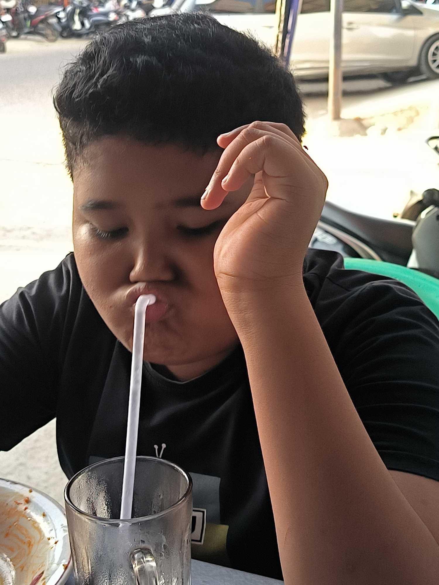 A boy is drinking from a glass with a straw while resting his head on his hand. He is wearing a black t-shirt. The scene appears to be outdoors in a public space, possibly a food stall or restaurant in Lhokseumawe, Indonesia. Motorbikes and a car are visible in the background, suggesting an urban street setting.