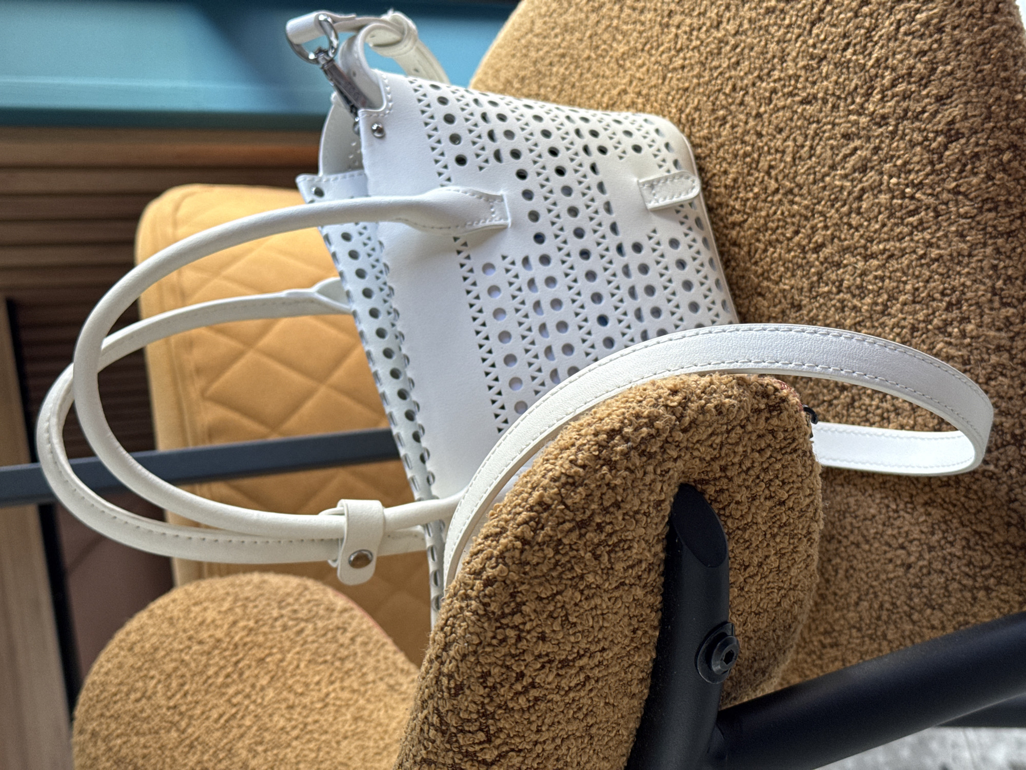 A white, perforated tote bag with two top handles and a detachable shoulder strap rests on a fuzzy, caramel-colored armchair. The bag is positioned slightly to the right of the frame, with its handles curving upwards. The armchair is situated in what appears to be a domestic or commercial indoor setting, with a bright yellow quilted chair visible in the background and a dark grey metal chair leg partially obscuring the left side of the image. The overall lighting suggests an indoor environment, possibly illuminated by natural light. The image captures a still life of the handbag and armchair, with no people or actions depicted. The location is described as Berdychiv, Ukraine.